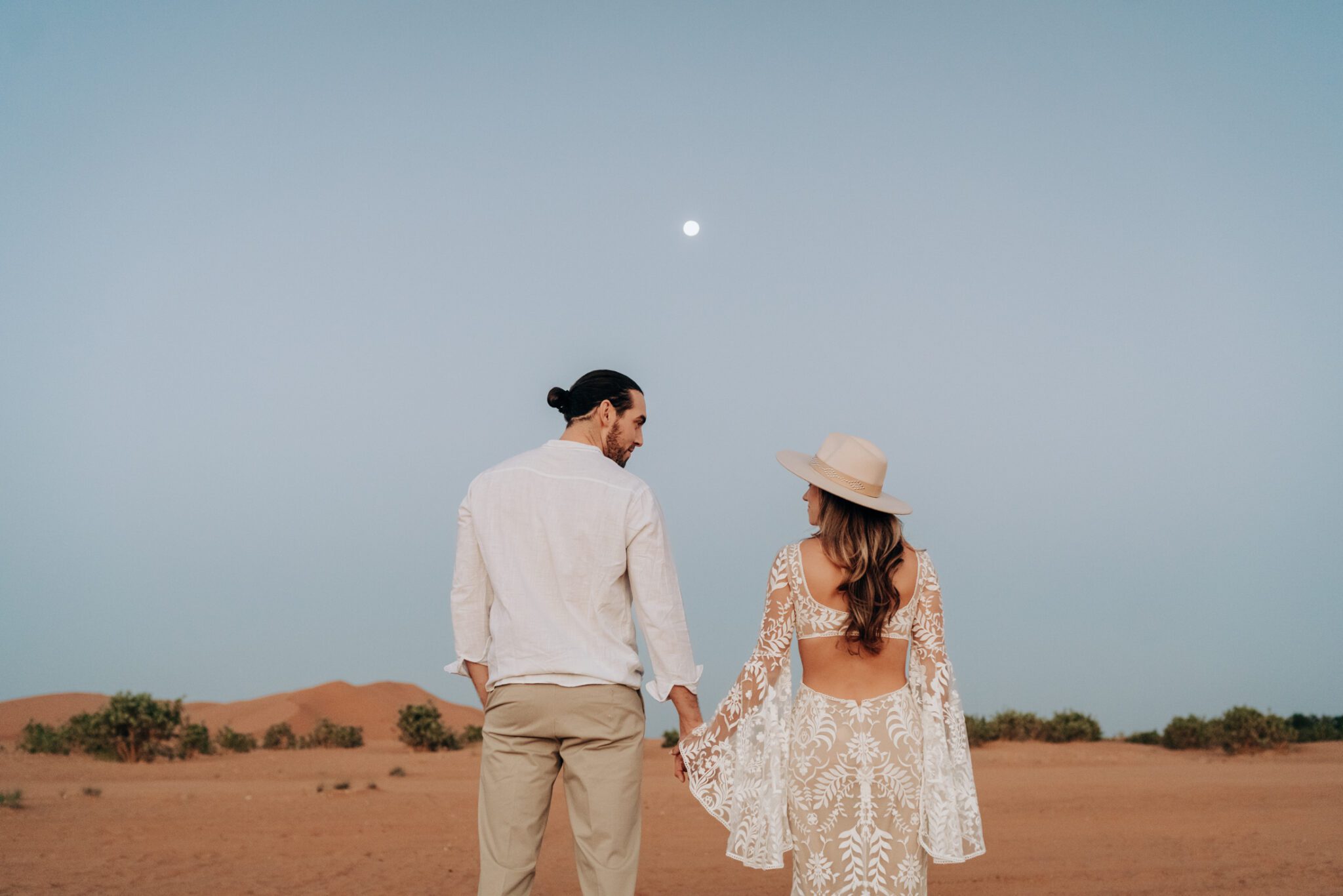 Couple holding hands in the desert with the moon in the background, captured by Seanna Leaf Photography.