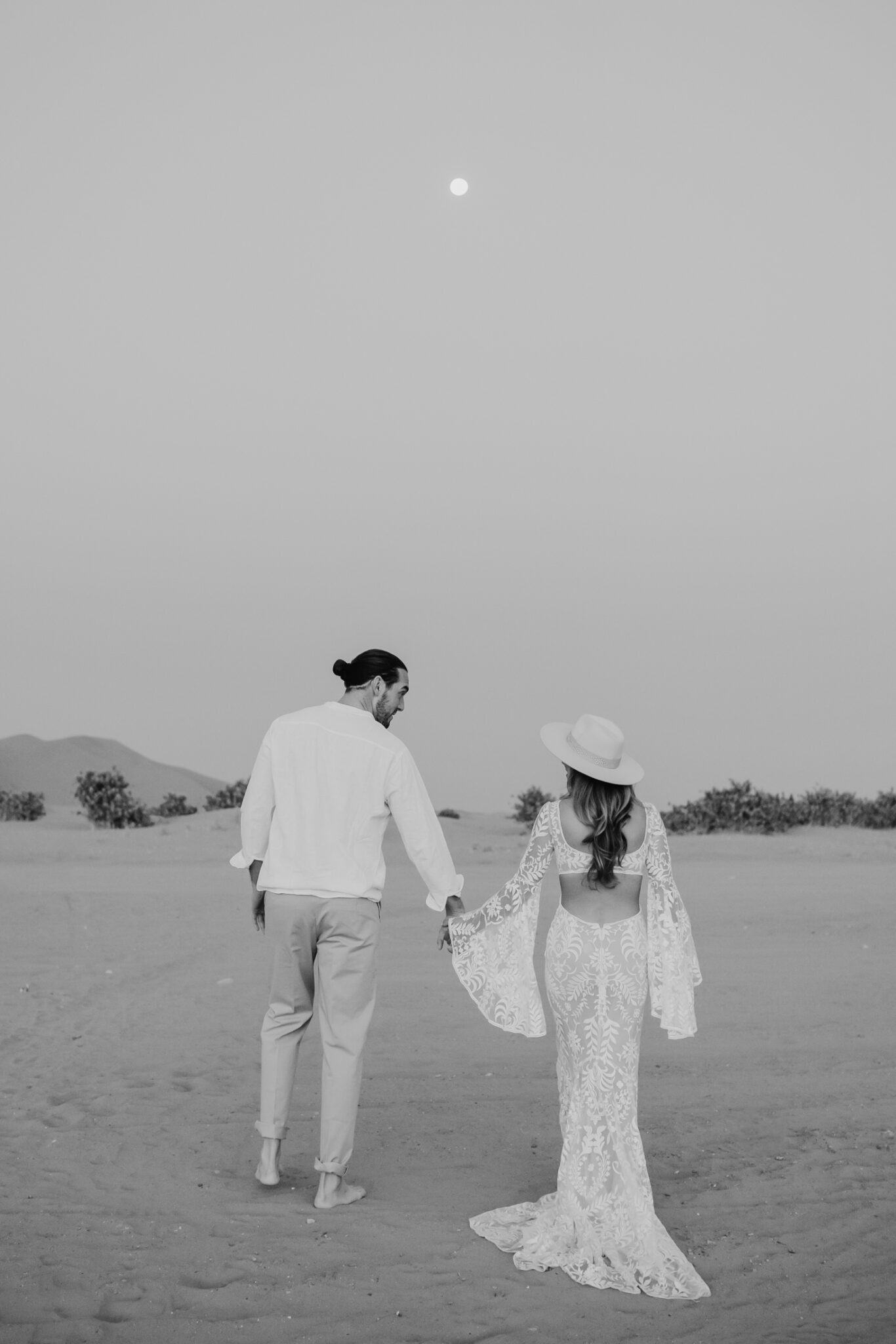 Couple holding hands walking through the desert, woman wearing boho inspired lace gown from The Wild Love Collective.