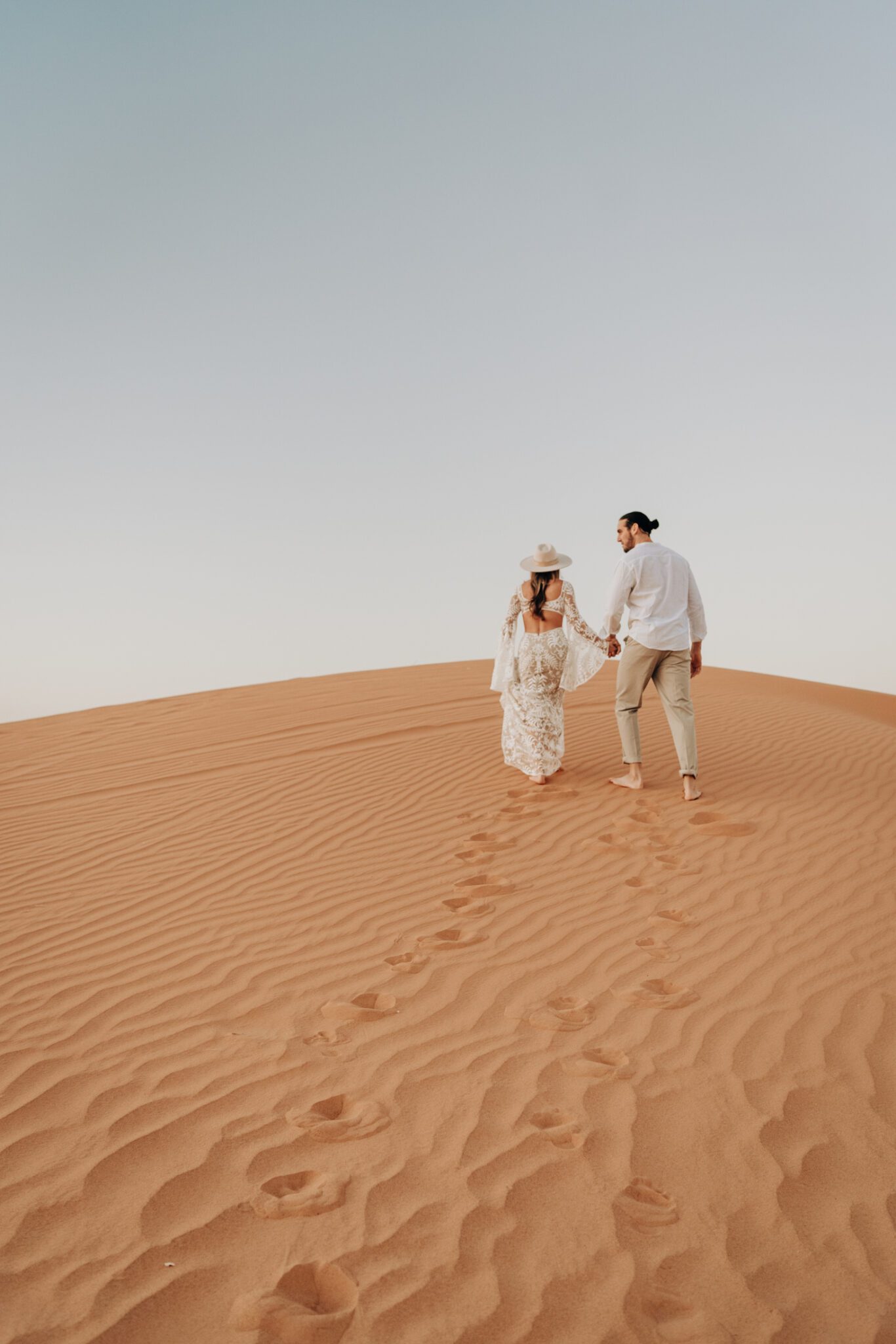 Couple holding hands walking through the sand dunes of a Moroccan desert, during their boho sunrise engagement session.