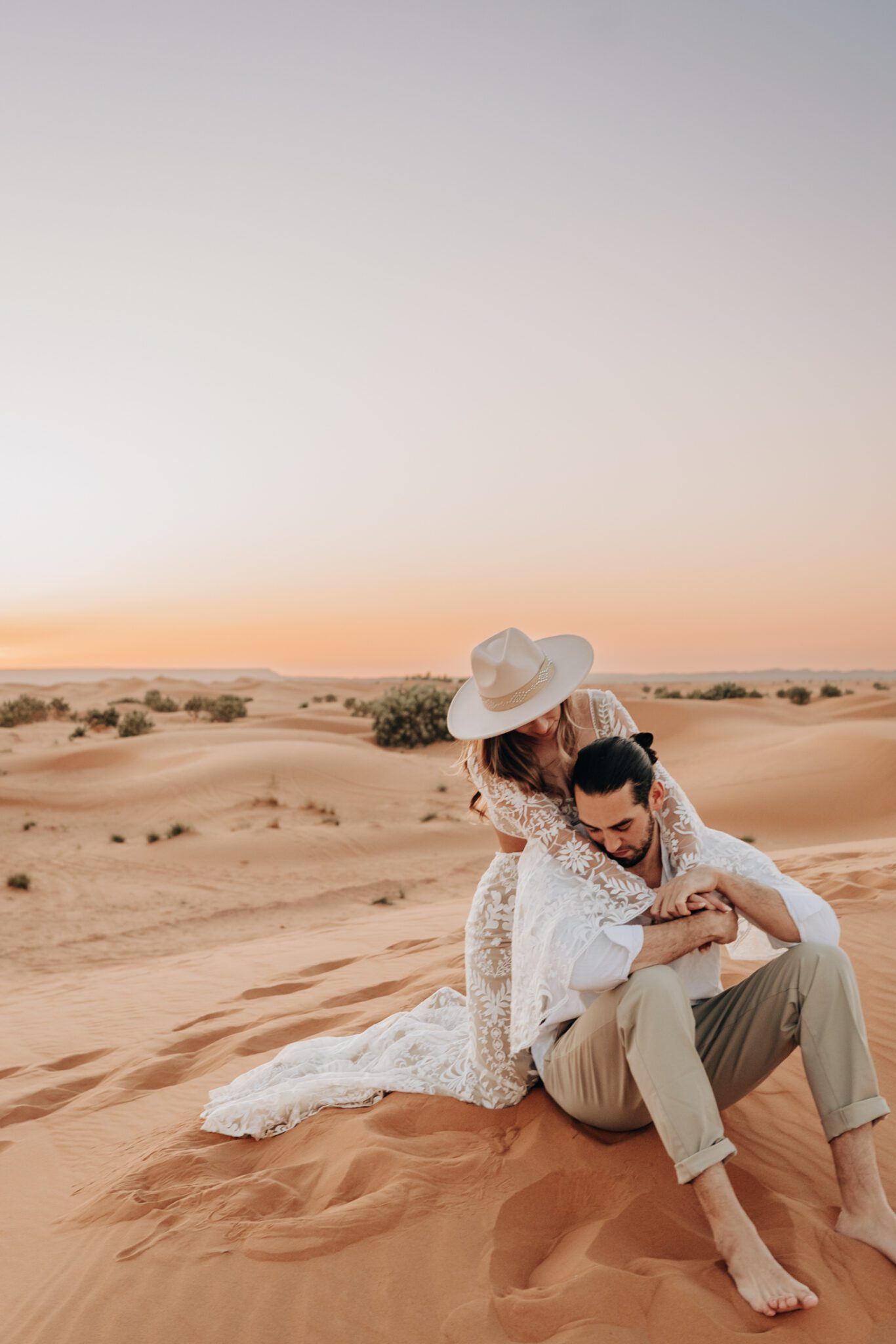 Couple sitting in the sand dunes of the Sahara Desert with golden sunrise in the distance, engagement session inspiration