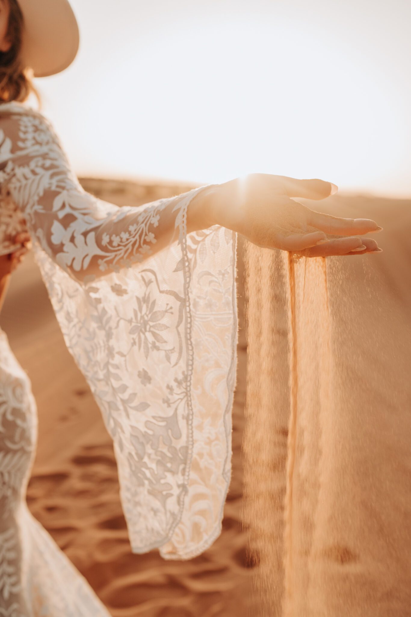 Bride playing with sand during Moroccan sunrise engagement session, wearing bohemian inspired lace gown with bell sleeves.