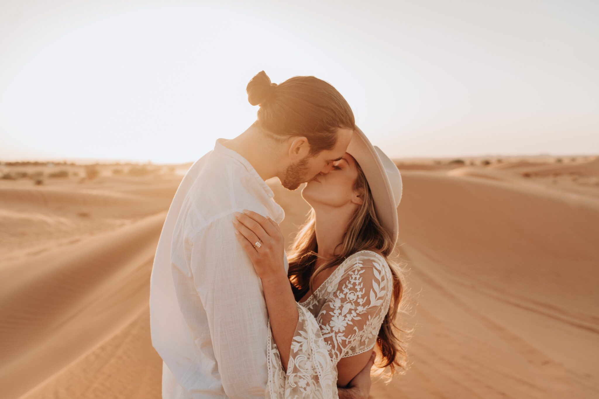 Couple kissing during their golden sunrise desert engagement session, woman wearing boho inspired lace gown.