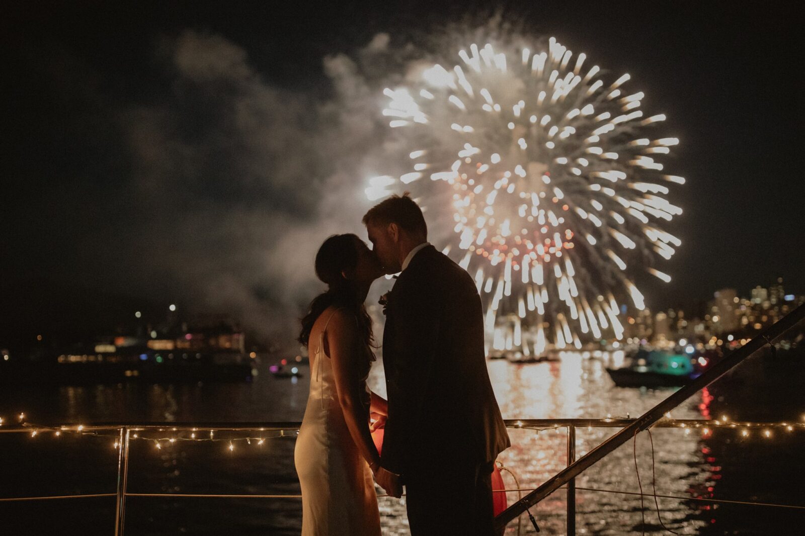 Bride and groom embrace on boat during Festival of Lights, watching fireworks on the ocean. 
