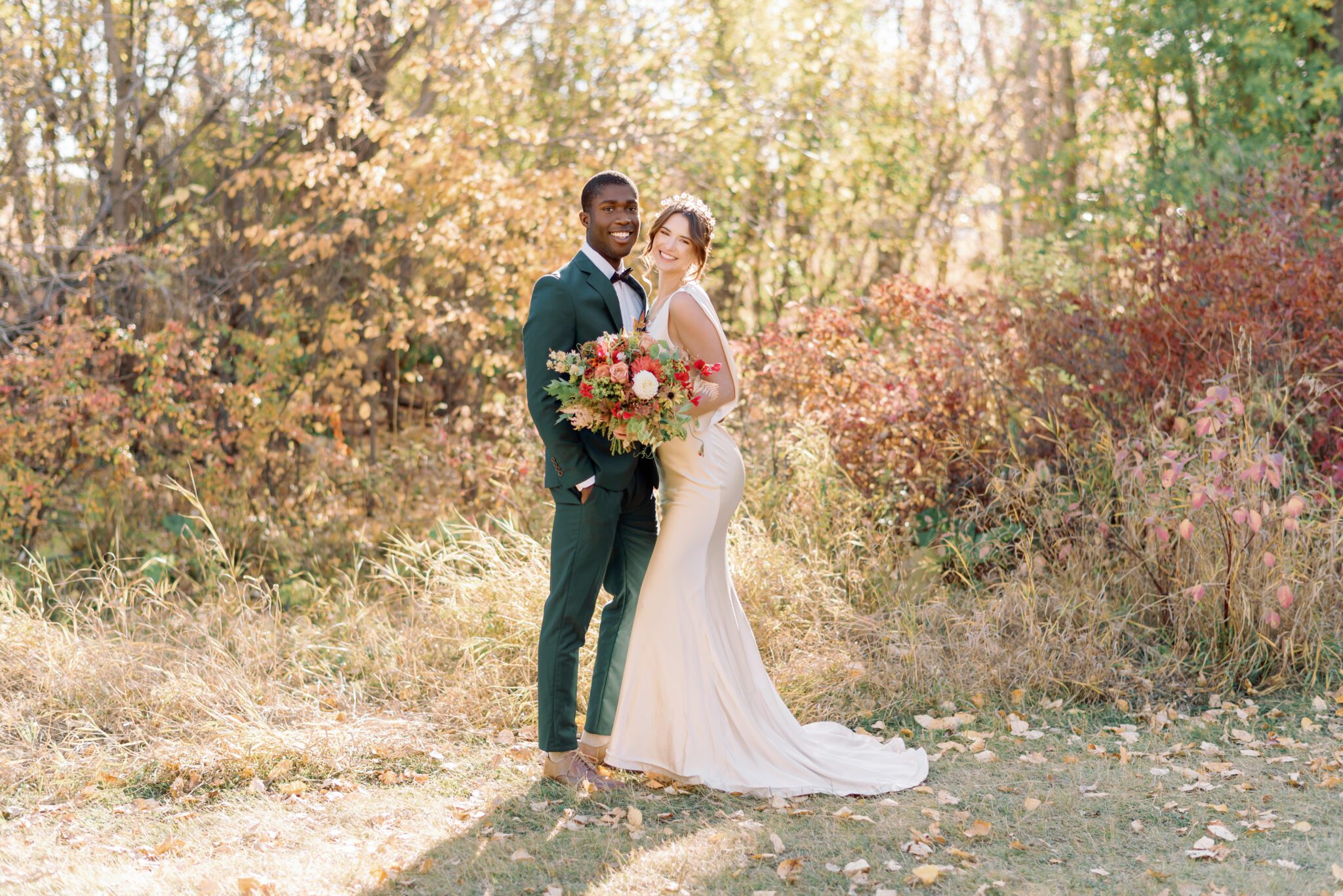 Couple embracing at intimate Fall wedding, groom wearing emerald green suit, and bride in elegant satin gown. Organic rich fall florals by Petal and Stem.