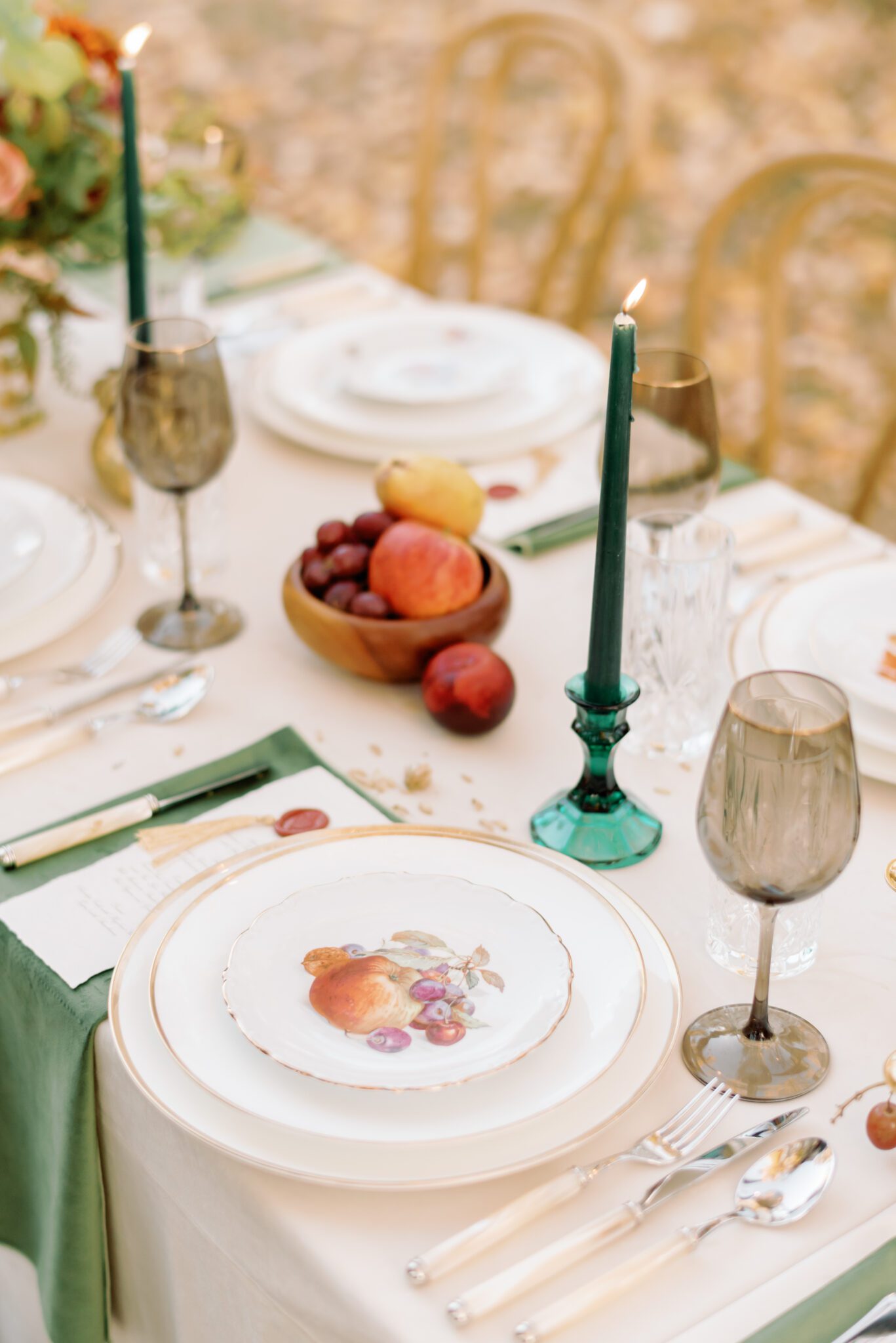 Al fresco dining at golden hour. Intimate autumn wedding reception tablescape featuring elegant menus with crimson wax seals and gold tassel accents, smoke tinted wine glasses with. agold rim, gold-rimmed vintage china with fruit paintings on them, pearl cutlery, and antique gold pear-shaped candleholders. Also includes olive green velvet napkins, wooden fruit bowl displays, and organic fall floral centerpieces. 