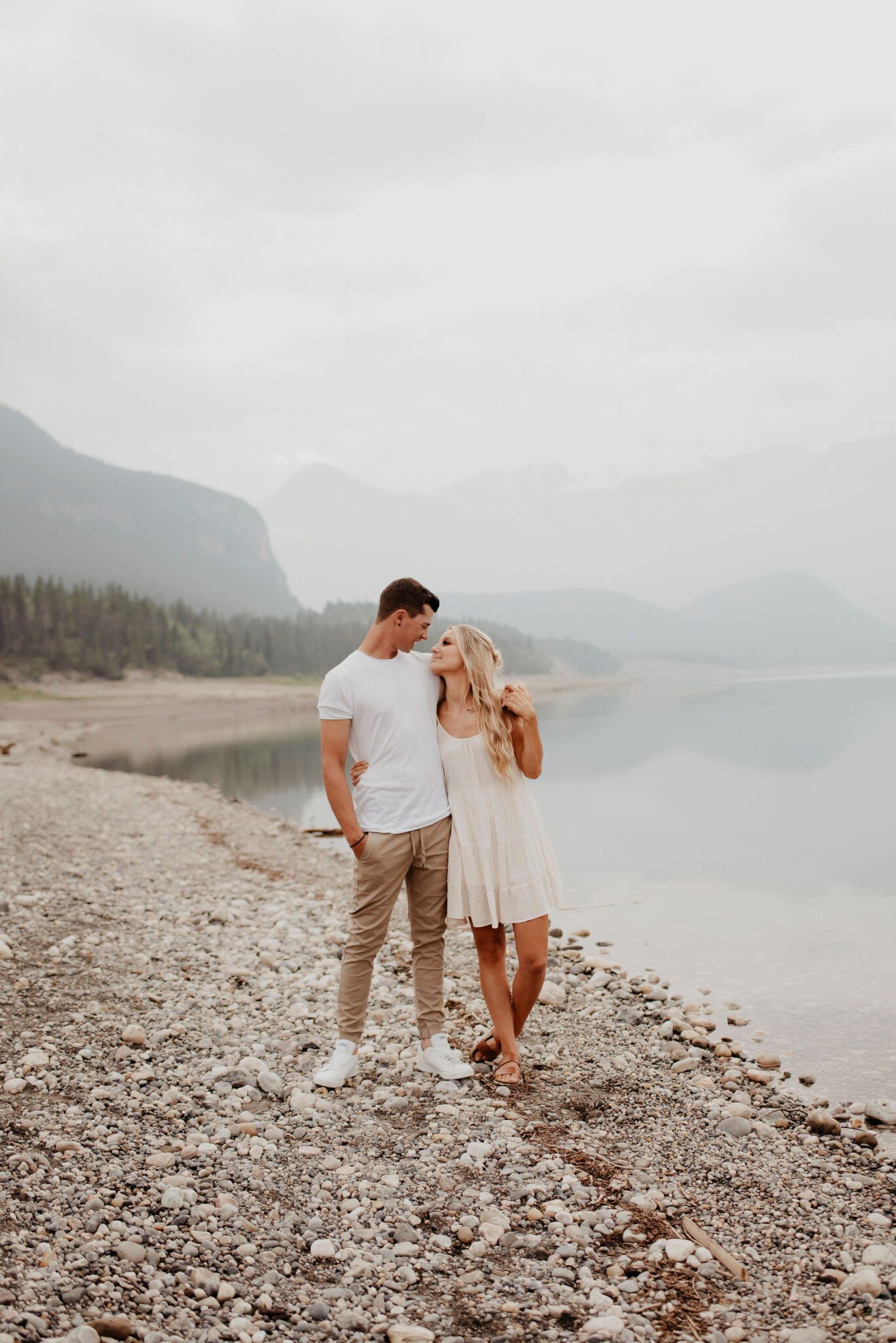 Couple standing on rock beach at Barrier Lake. Summer engagement session outfit ideas, woman wearing flowy white linen dress, and man wearing white tee-shirt and tan jogger panes with white sneakers. 