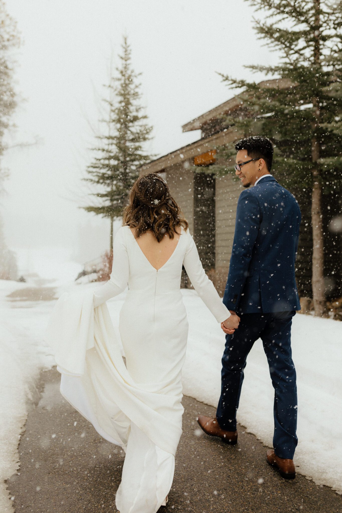 Snow is falling at the Silvertip Resort as the newly married couple walks along a snow-covered path, bride is wearing elegant gown, winter wedding inspiration. 