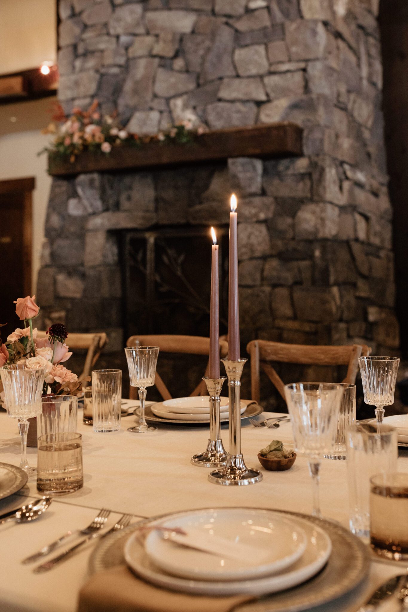 Warm wood tones and rich colours at this Silvertip Resort wedding reception, with a warm-toned tablescape, vintage glassware, and a large stone fireplace as the backdrop, winter wedding inspiration. 