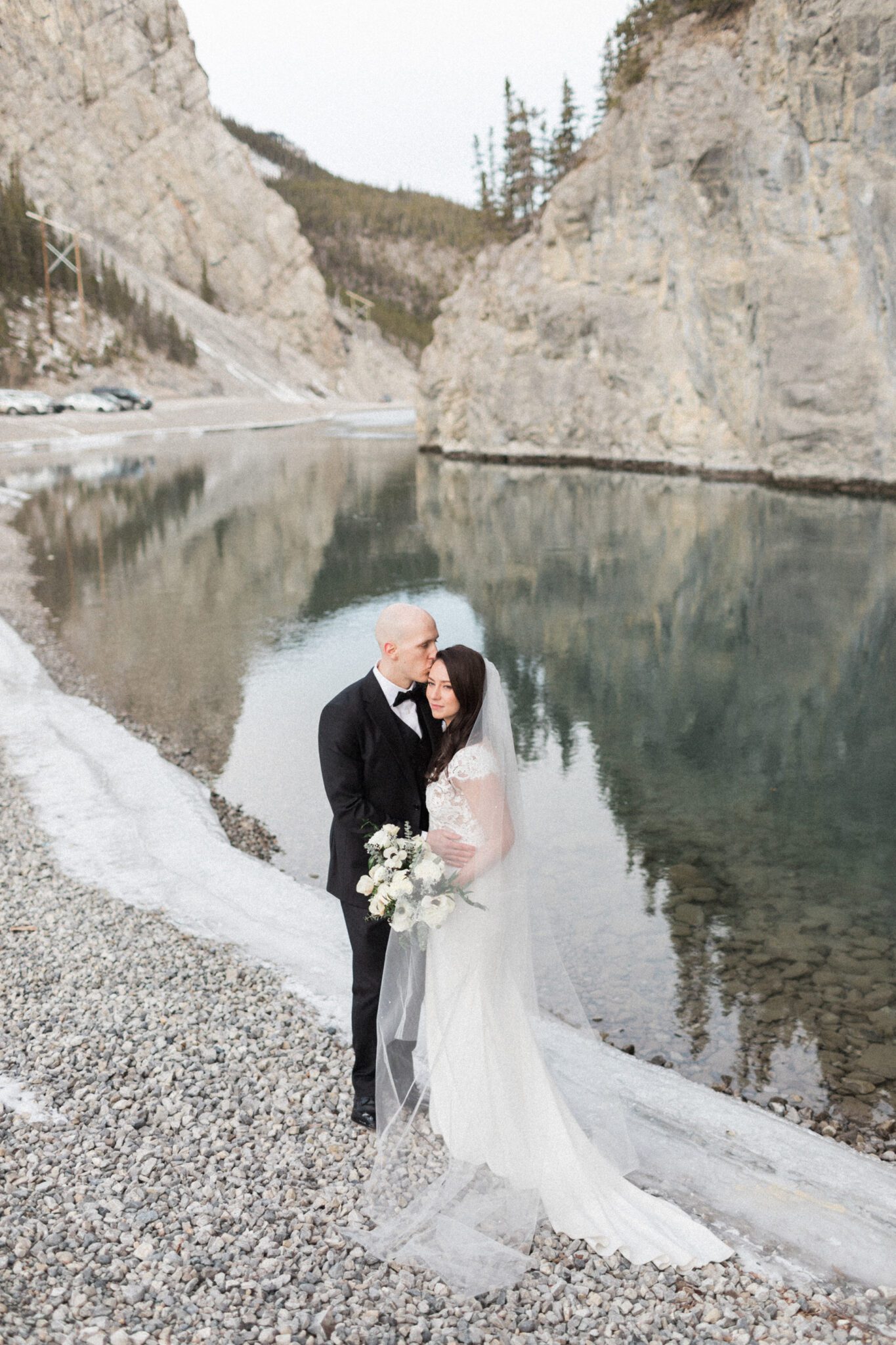 Portrait of bride and groom in front of mirror lake with a gorgeous mountain backdrop, bride holding elegant bouquet white white florals and greenery. Winter wedding inspiration. 