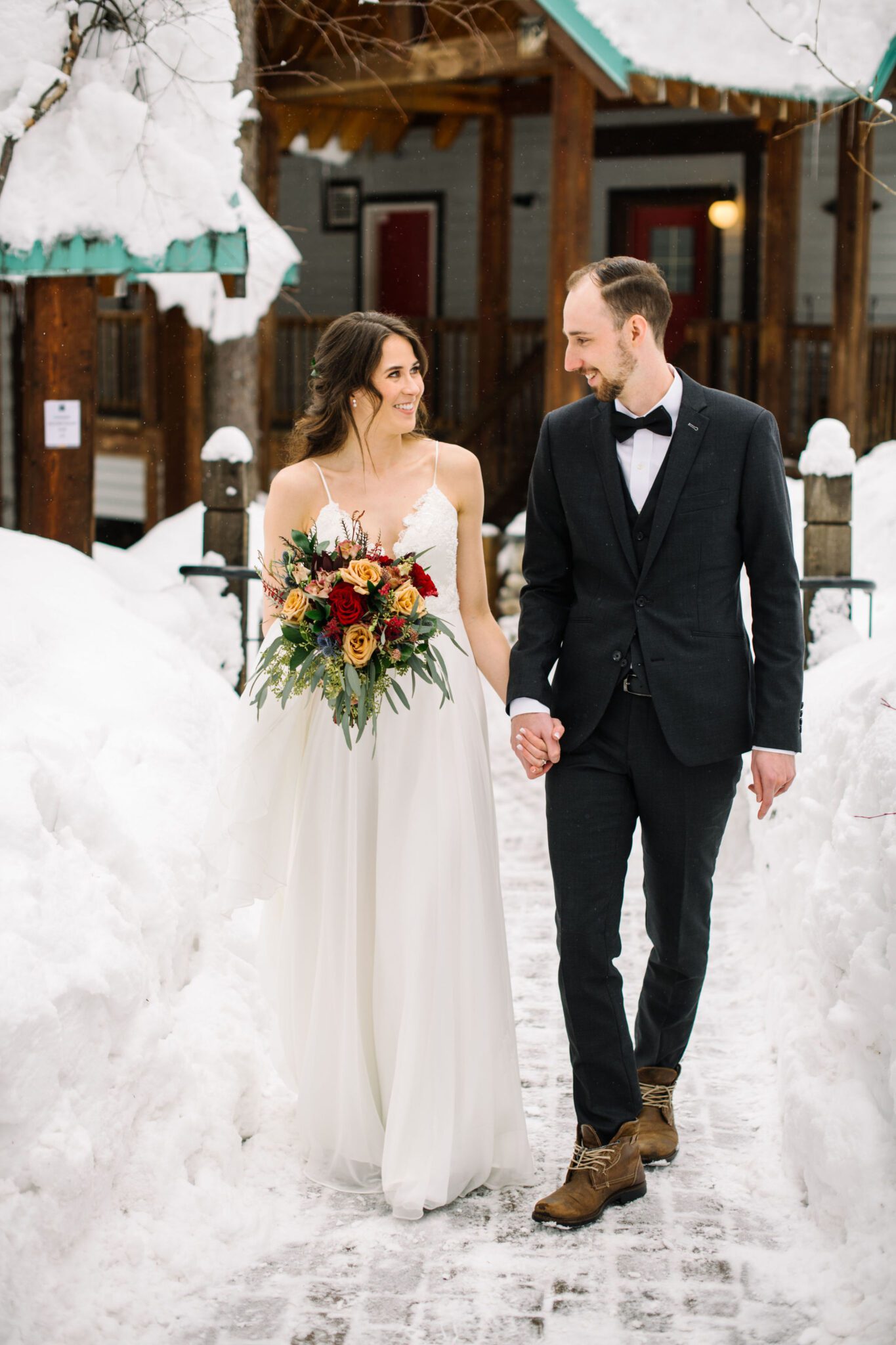 Portrait of couple in front of snow-covered cabin at Emerald Lake Lodge, bride holding winter-themed bouquet, winter wedding inspiration. 