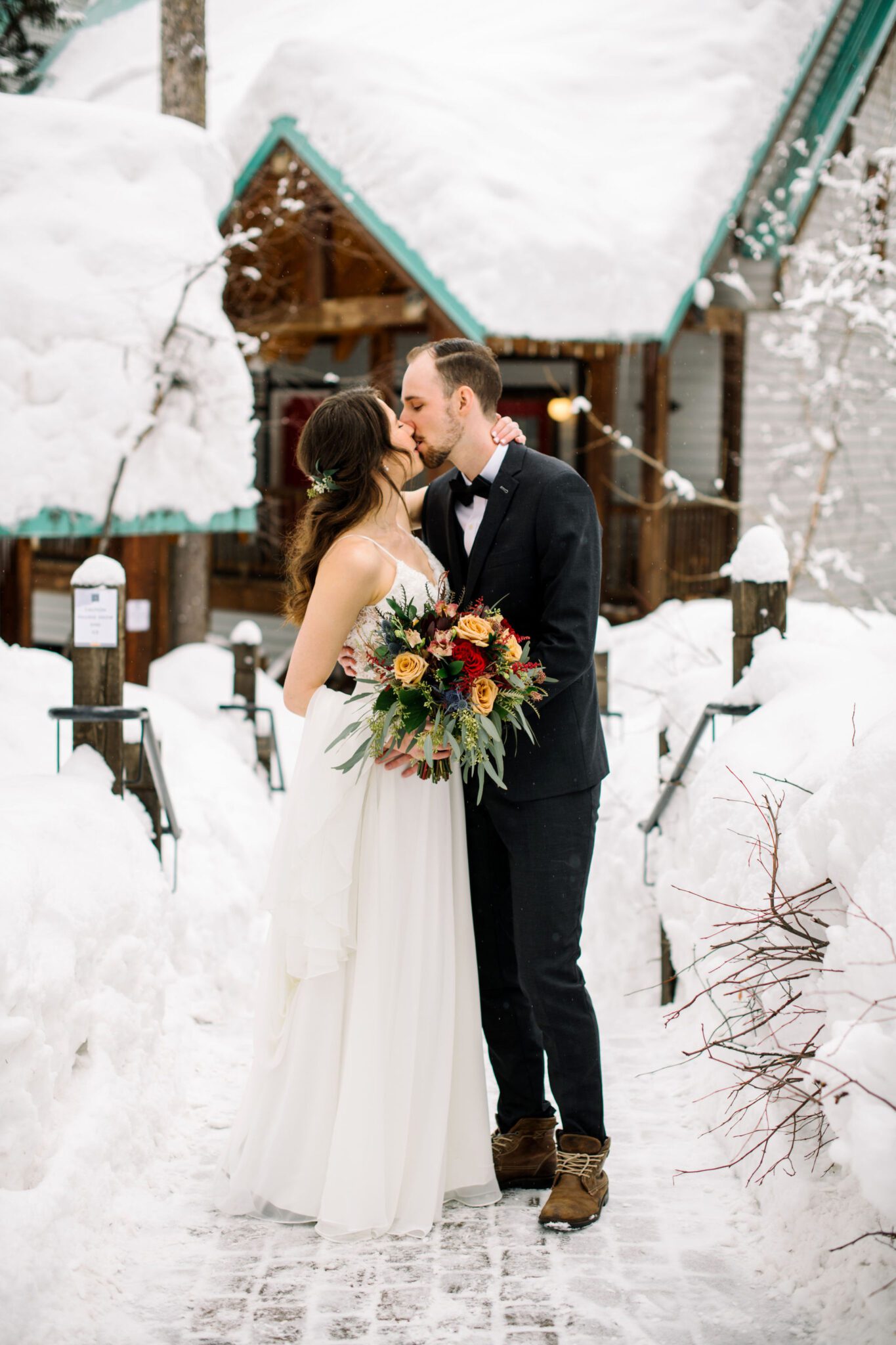 Portrait of couple kissing in front of cozy cabin at Emerald Lake Lodge in Alberta, bride holding bouquet with winter-themed colours, winter wedding inspiration. 