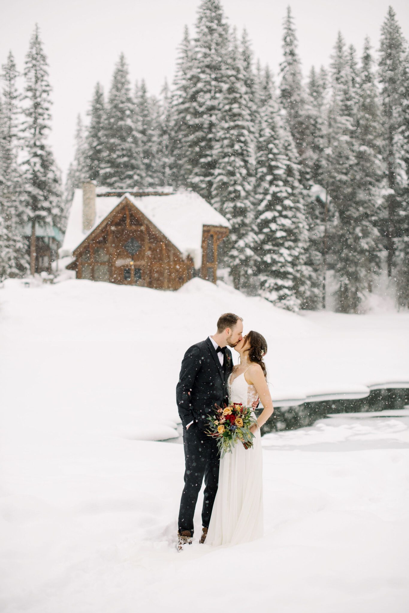 Wide portrait of couple sharing a kiss in front of the main lodge at Emerald Lake, covered in snow, winter wedding inspiration. 