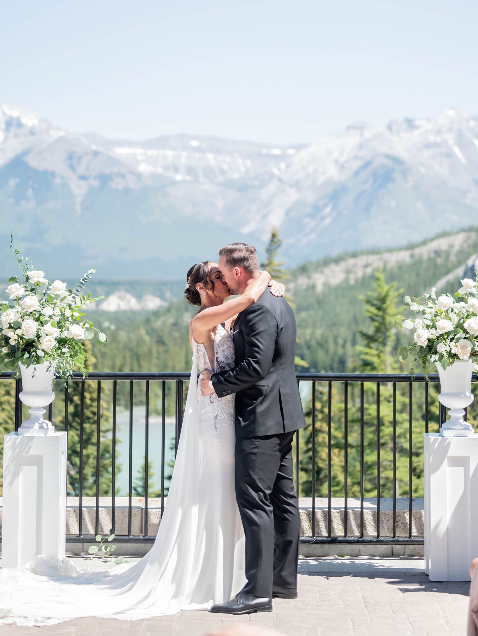 Bride and groom share first kiss at their Banff summer wedding surrounded by pedestals of white florals and greenery, classic wedding style inspiration. 