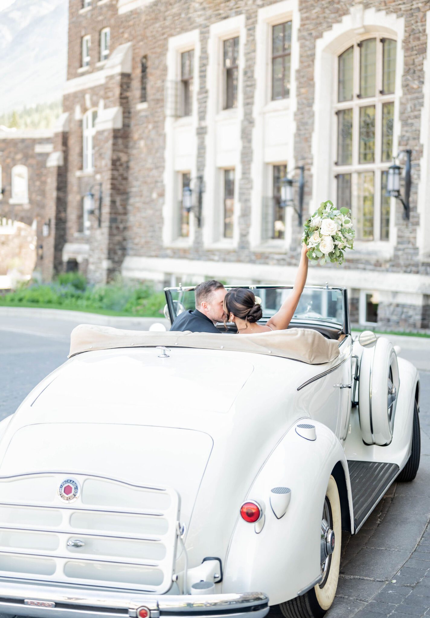 Bride and groom exiting wedding ceremony in cream-coloured vintage car, white floral bouquet in the air at the Fairmont Banff Springs. 