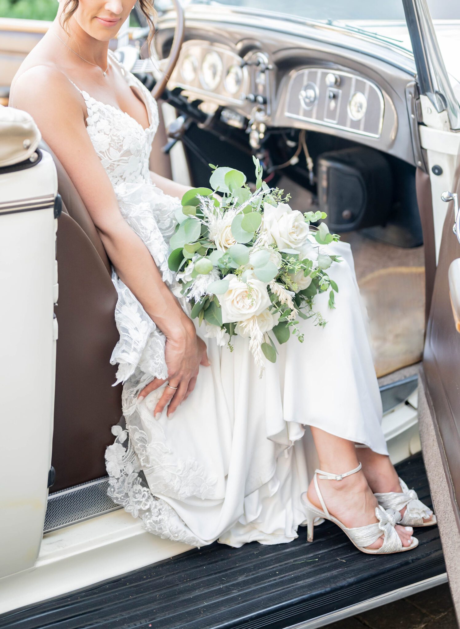 Bride leaning on the side of vintage car in elegant lace gown, holding white florals with greenery at Banff Summer Wedding. 