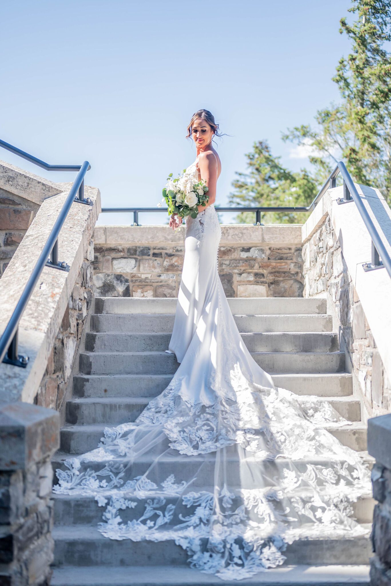 Elegant portrait in the summer sun of bride on the staircase of the Fairmont Banff Springs, lace wedding gown draping down, bride holding white floral bouquet with greenery. 