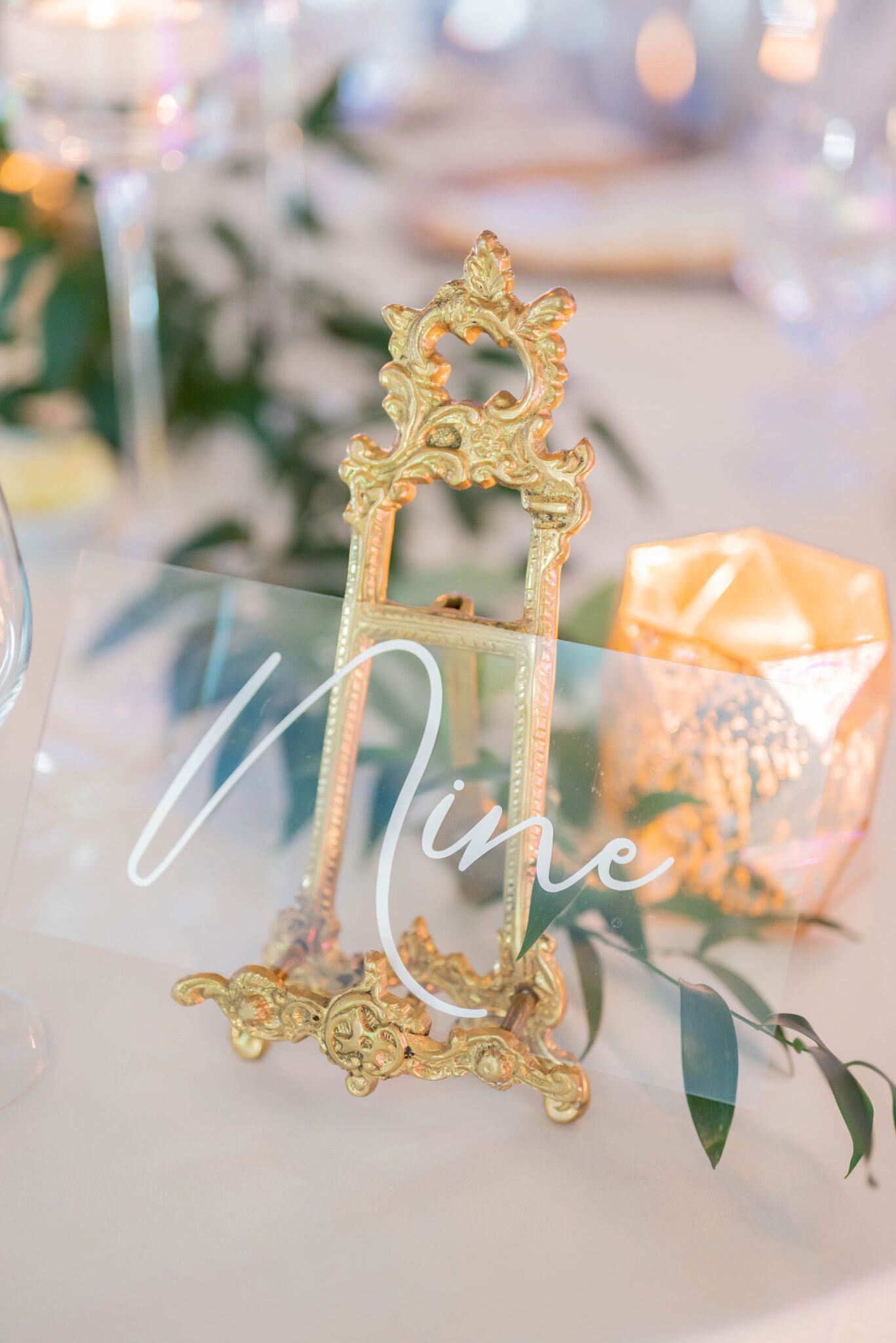 Hand-painted table numbers with gold detailed stand, surrounded by greenery, white linens, and gold candle holders, classic wedding tablescape inspiration