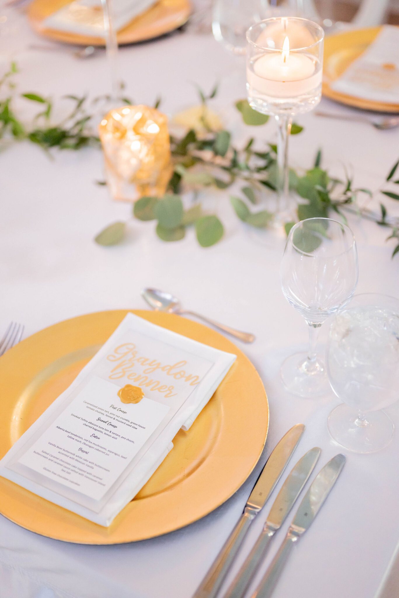 Classic wedding style with gold charger plates, custom menus for guests with gold handwriting, votive candles, and light greenery along the table. 