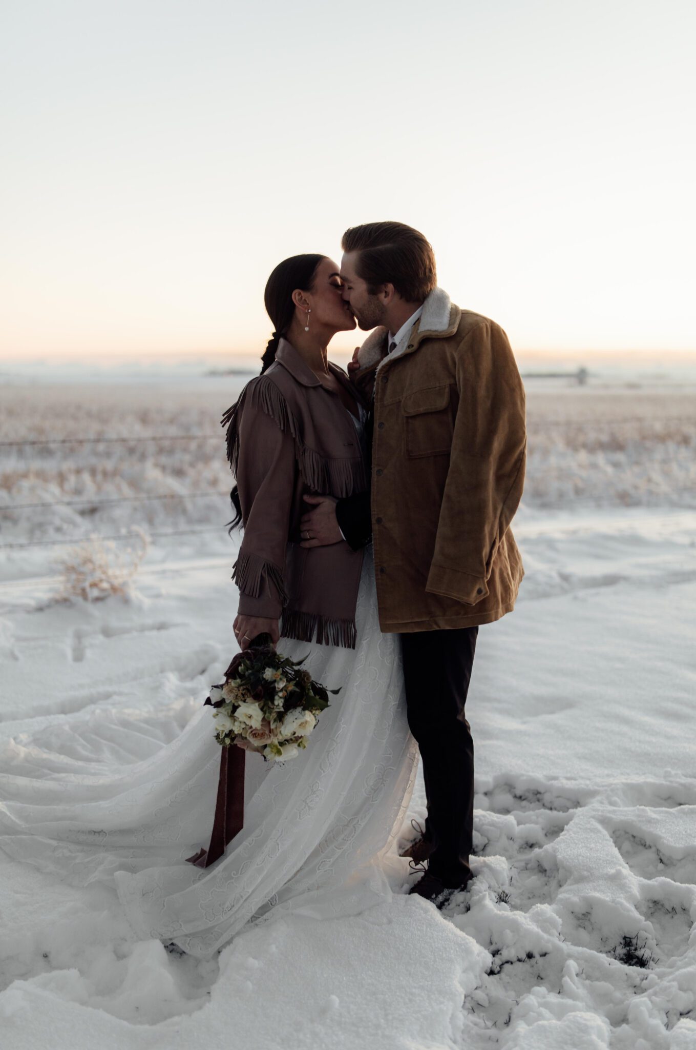 Wedding Inspiration With An Updated Take on Country Chic Wedding Style at Countryside Barn in Lethbridge Alberta, portrait of bride and groom kissing at sunset, winter wedding inspiration, groom's vintage bolo tie and thrifted suede fringe jacket from Alice & Irene. 