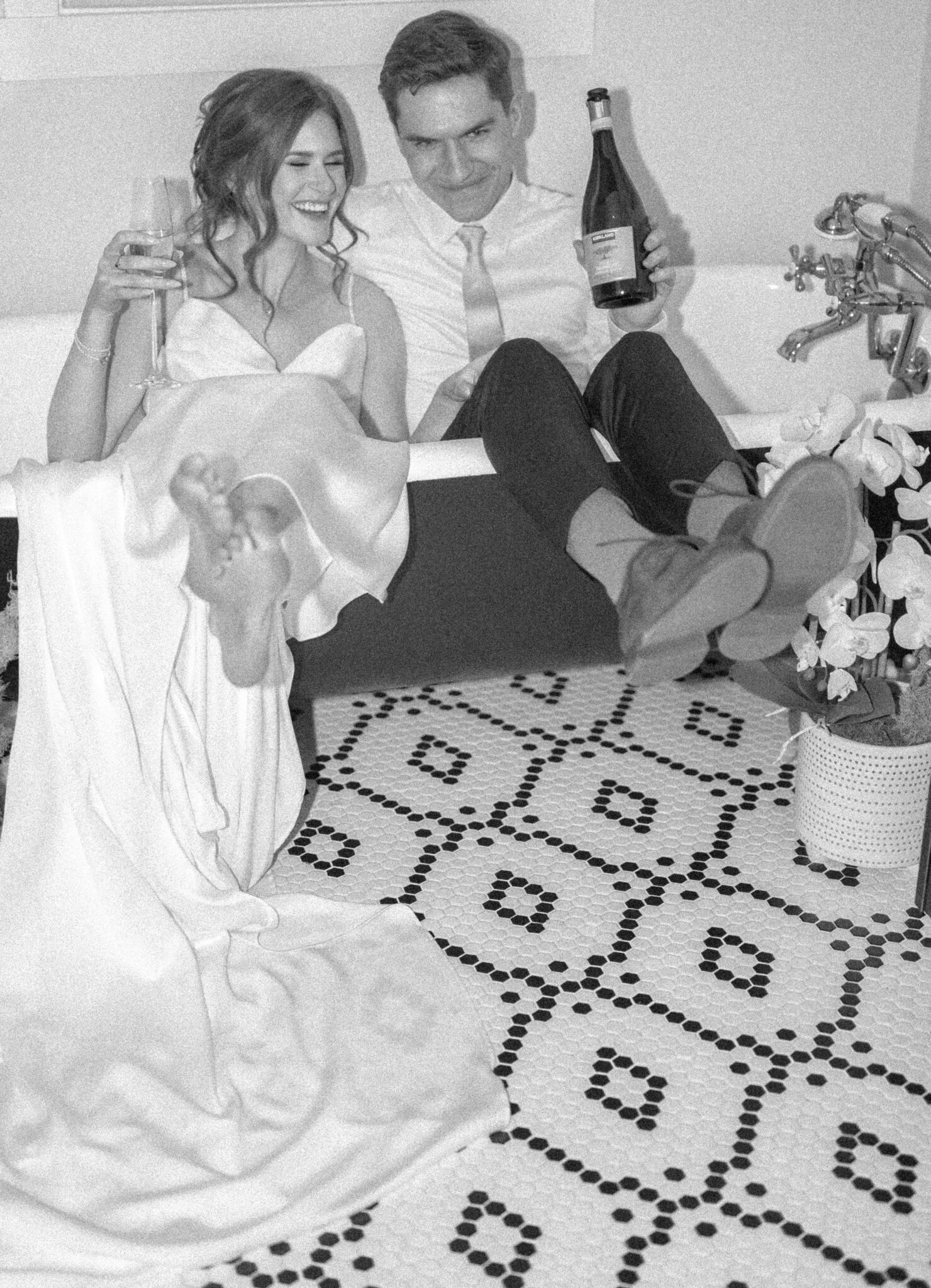 Couple shares a bottle of champagne in a clawfoot bathtub in the charming bridal suite with textured black and white tile, flash wedding portrait inspiration. 