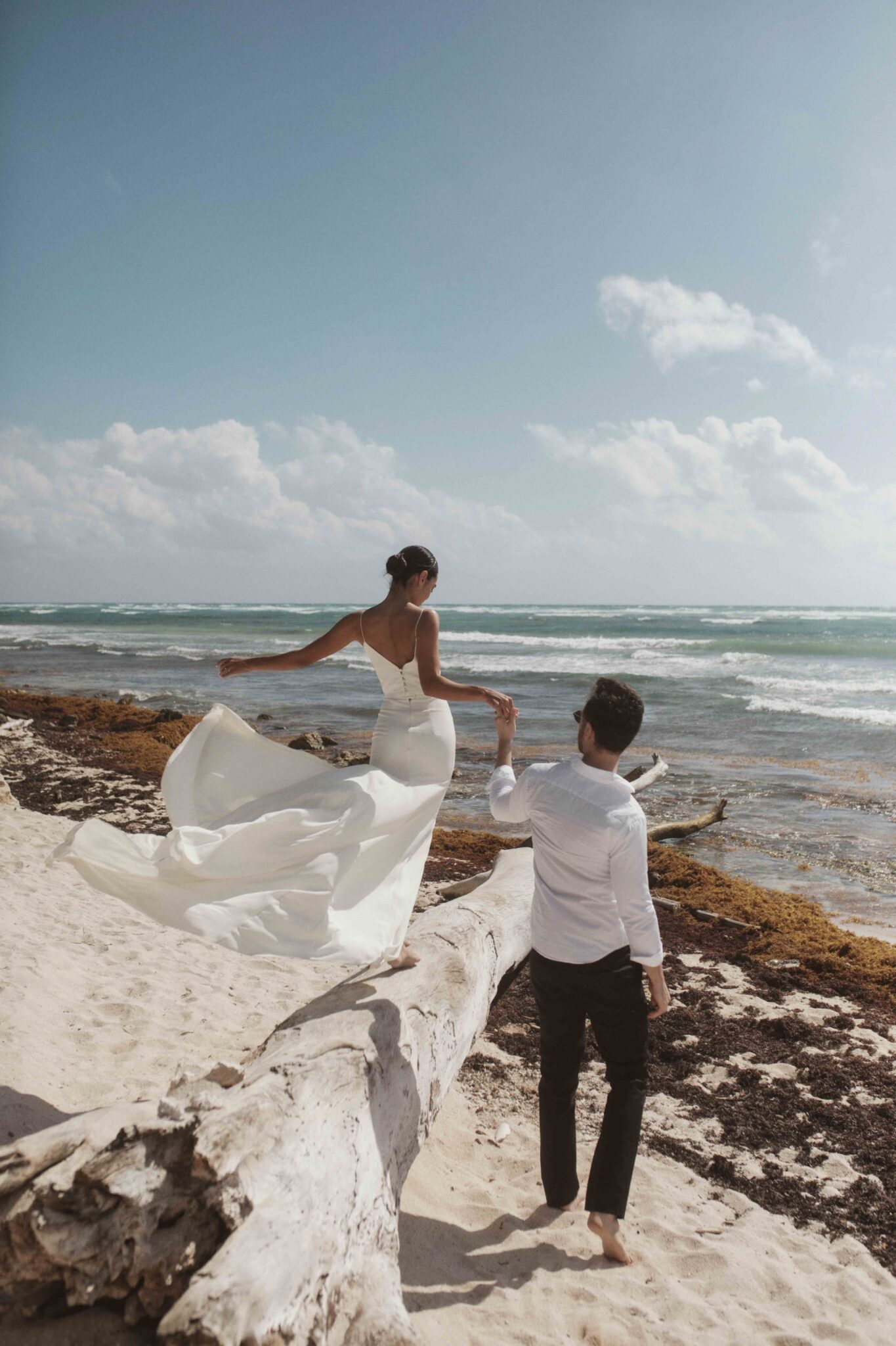 Seaside wedding portraits on the sandy shores of Mexico, groom leading bride along the shore, Canadian bridal designer Tempête's contemporary collection.