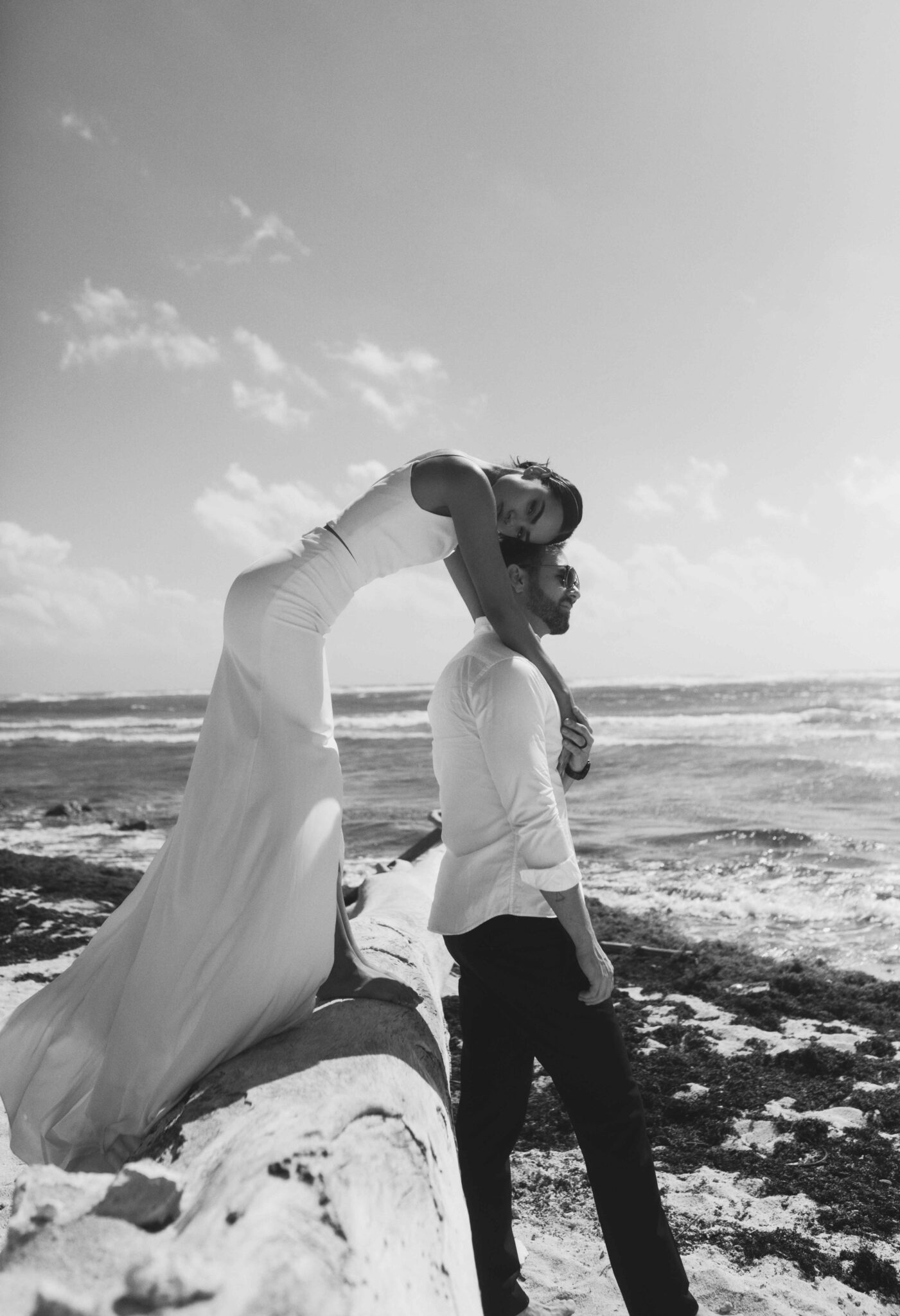 Seaside portrait of bride in elegant gown leaning over groom, as he gazes out onto the ocean, black and white portrait inspiration. 