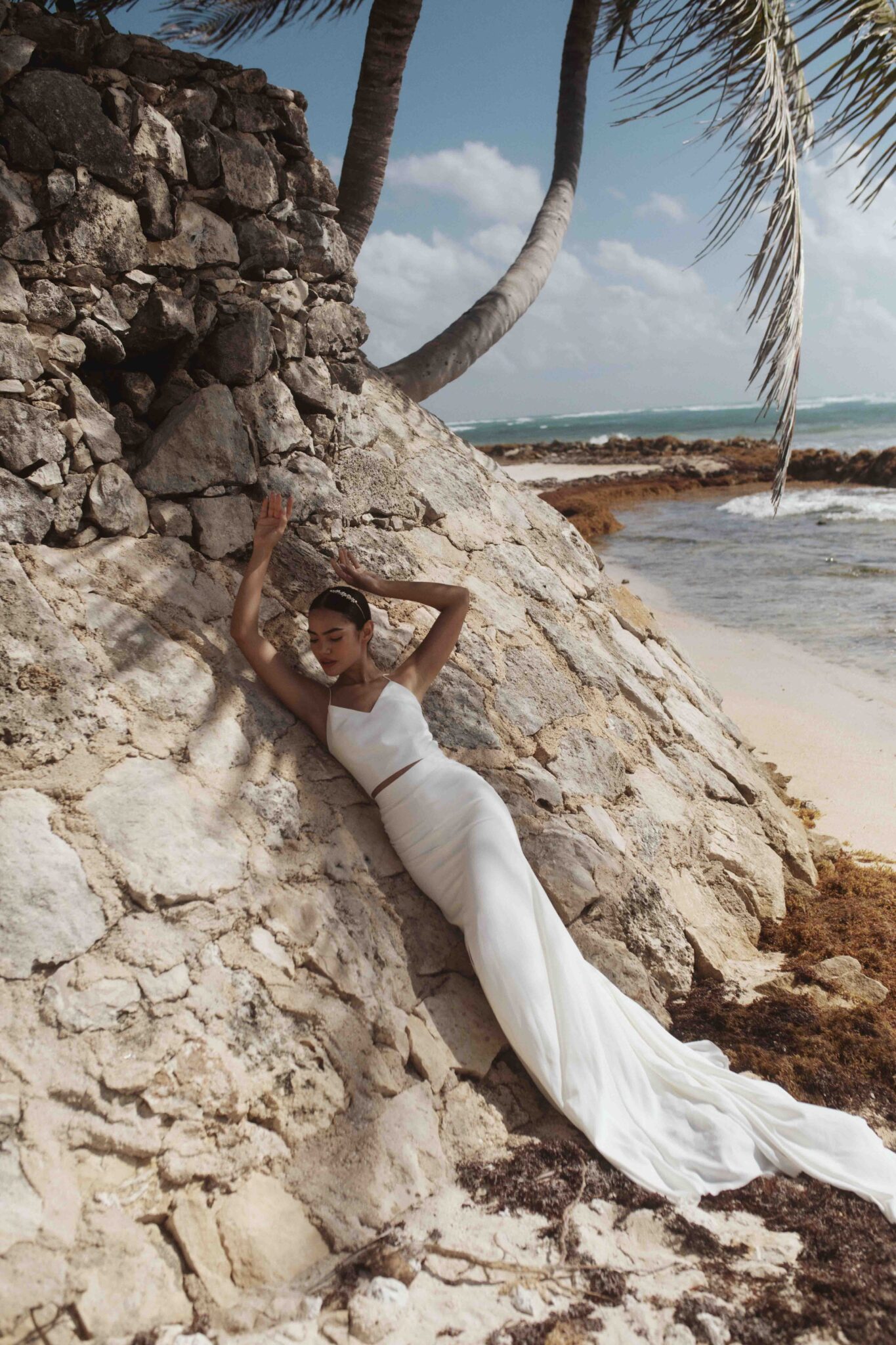 Seaside portrait of bride in TEMPÊTE chic gown, leaning against rock, Storm-inspired bridal inspiration from Tempête with an effortless & free-spirited nature. 