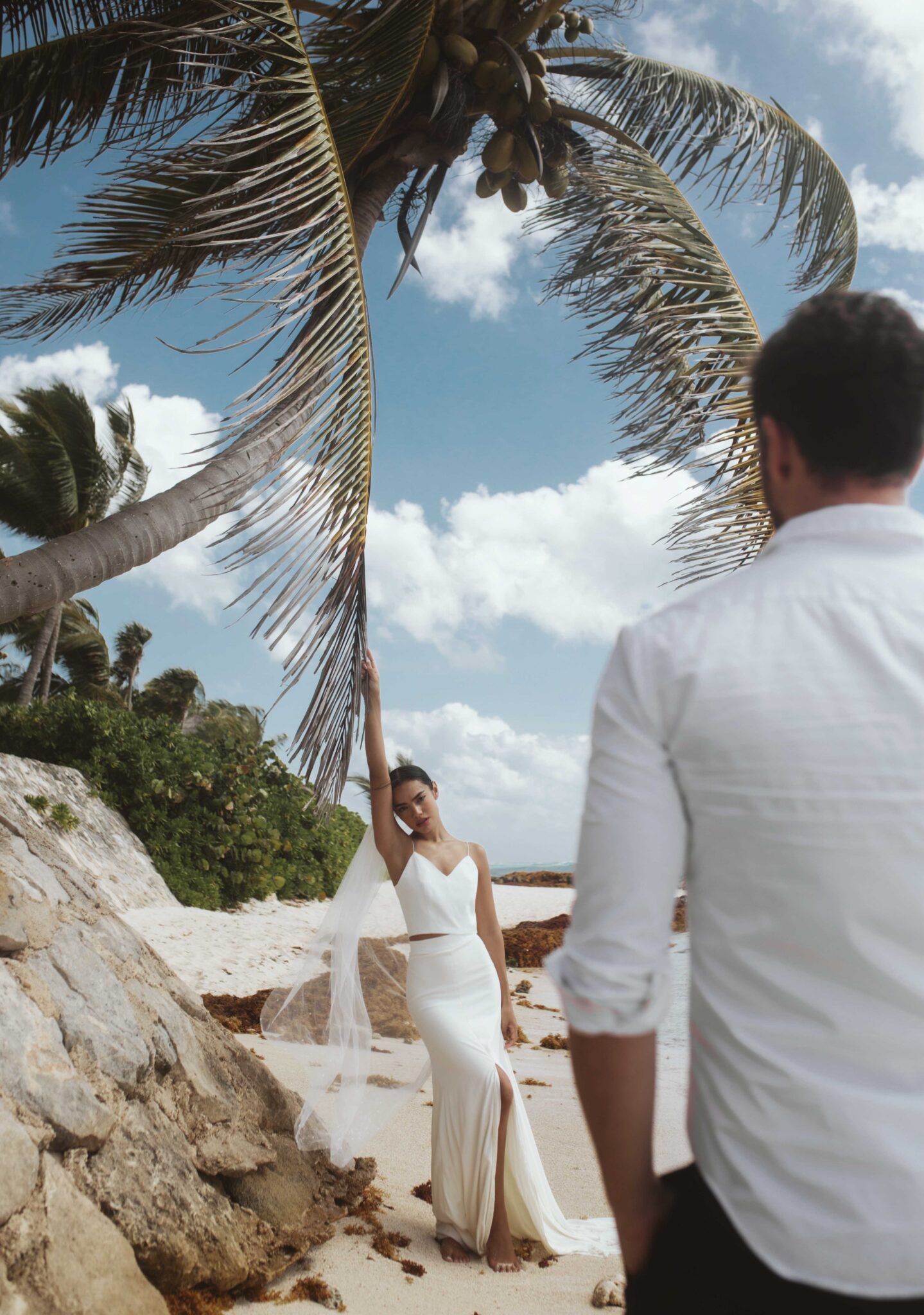 Groom gazing at his bride in Mexico on the sandy shores, while bride tugs from the palm tree, Storm-inspired bridal inspiration from Tempête with an effortless & free-spirited nature. 