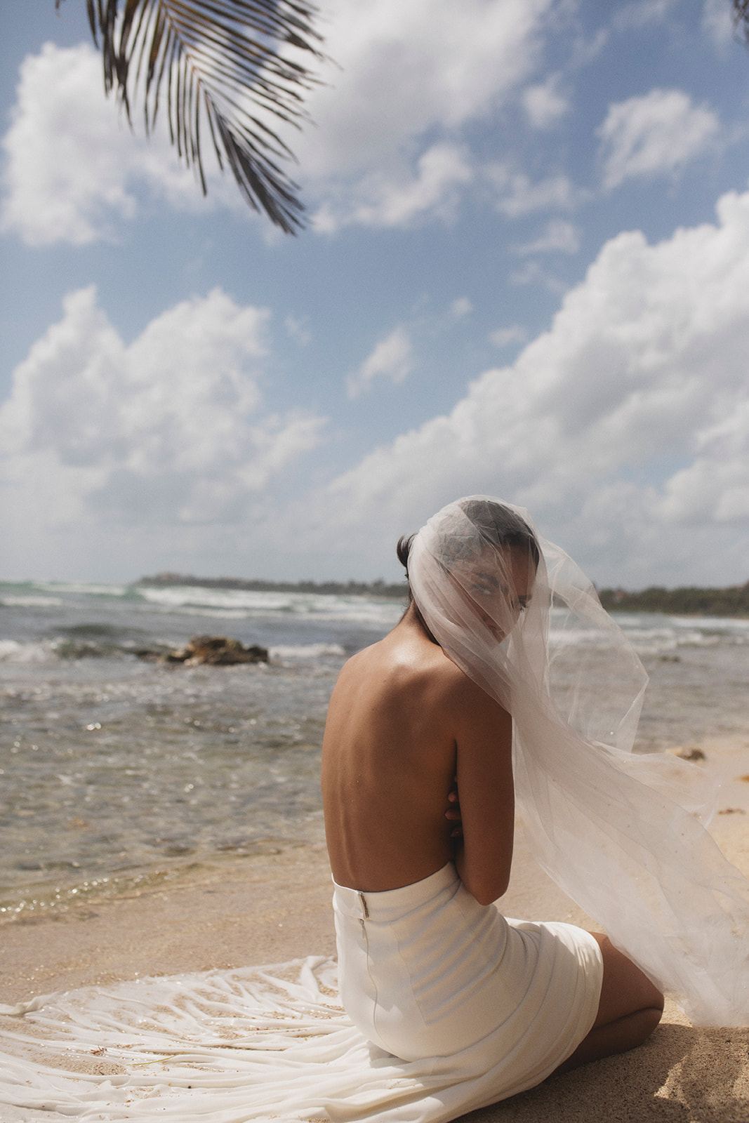 Bride sitting on the sandy shores, while the veil is blowing across her face, Storm-inspired bridal inspiration from Tempête with an effortless & free-spirited nature. 