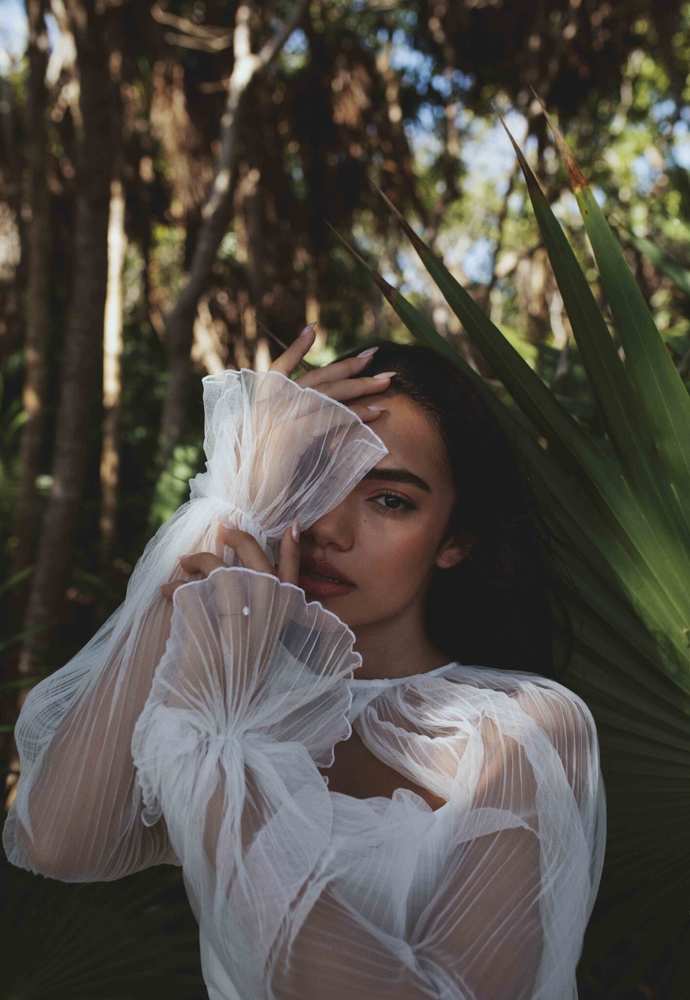 Close up portrait of bride holding arms close to her face, with elegant tulle sleeves, palm trees as the backdrop. 
