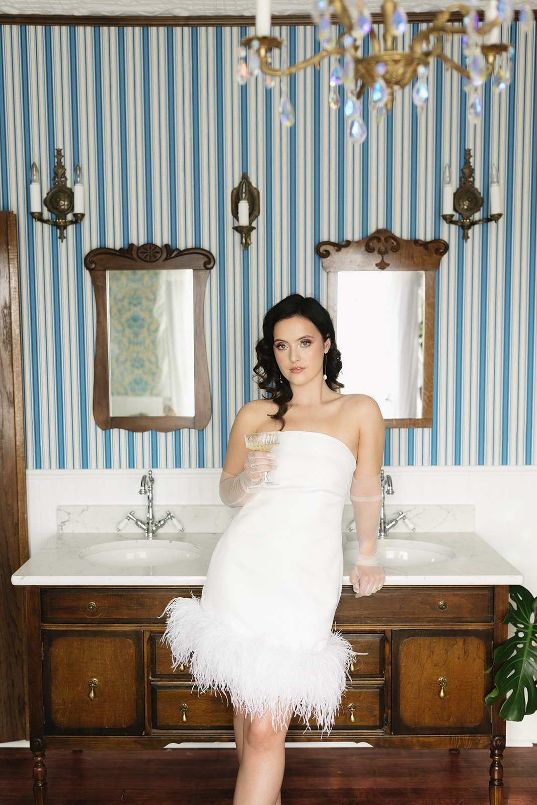 Fashion-forward bride leaning on bathroom counter wearing trendy feather-trimmed bridal gown. Morning-of Bridal Inspiration with Modern Style & Chic Getting Ready Photos.