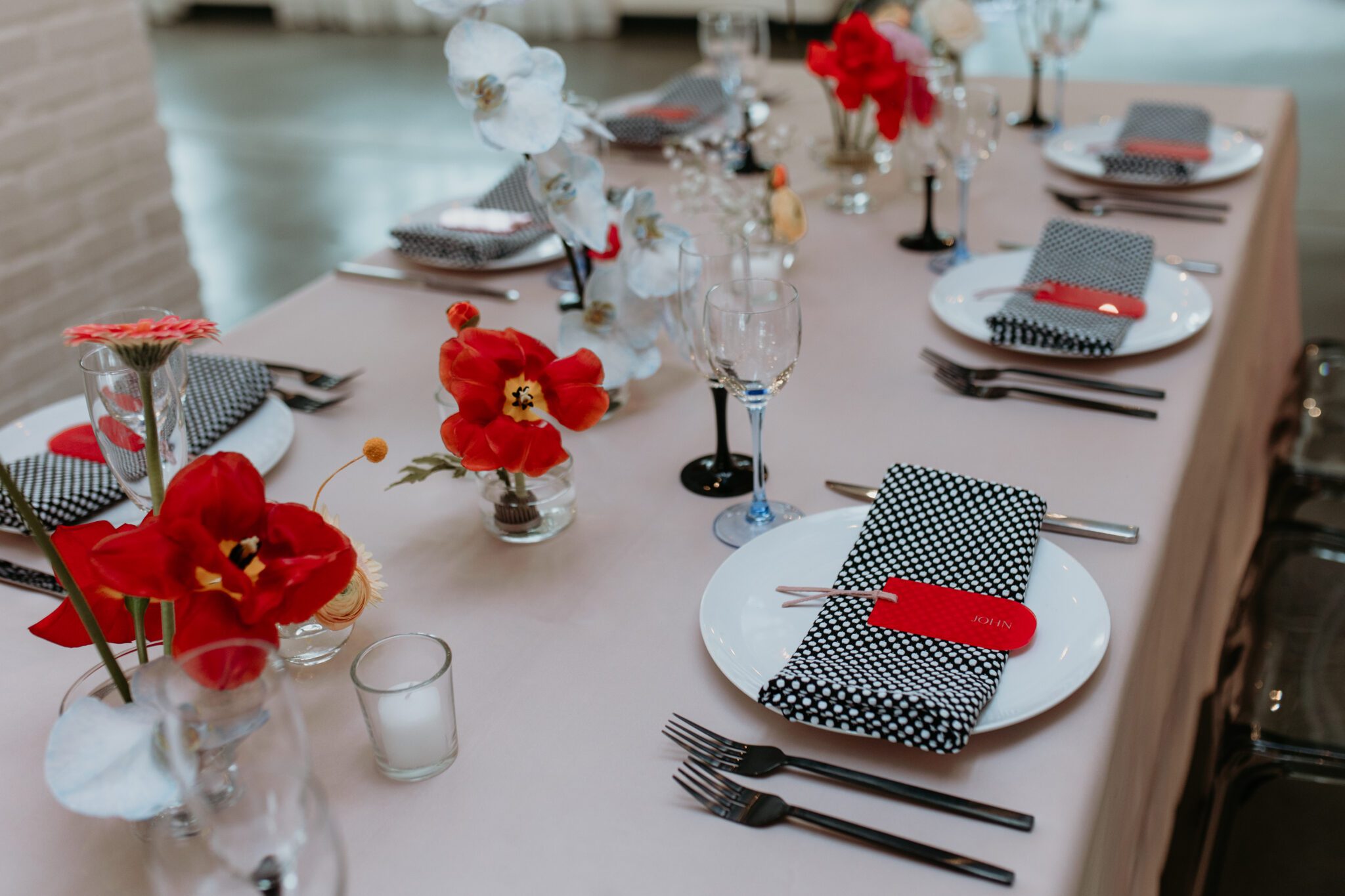 A contemporary wedding tablescape blending modern and retro elements: light pink tablecloth, grey ghost chairs, black acrylic place cards, patterned napkins, retro-inspired table numbers, and ikebana floral arrangements with anthurium flowers.