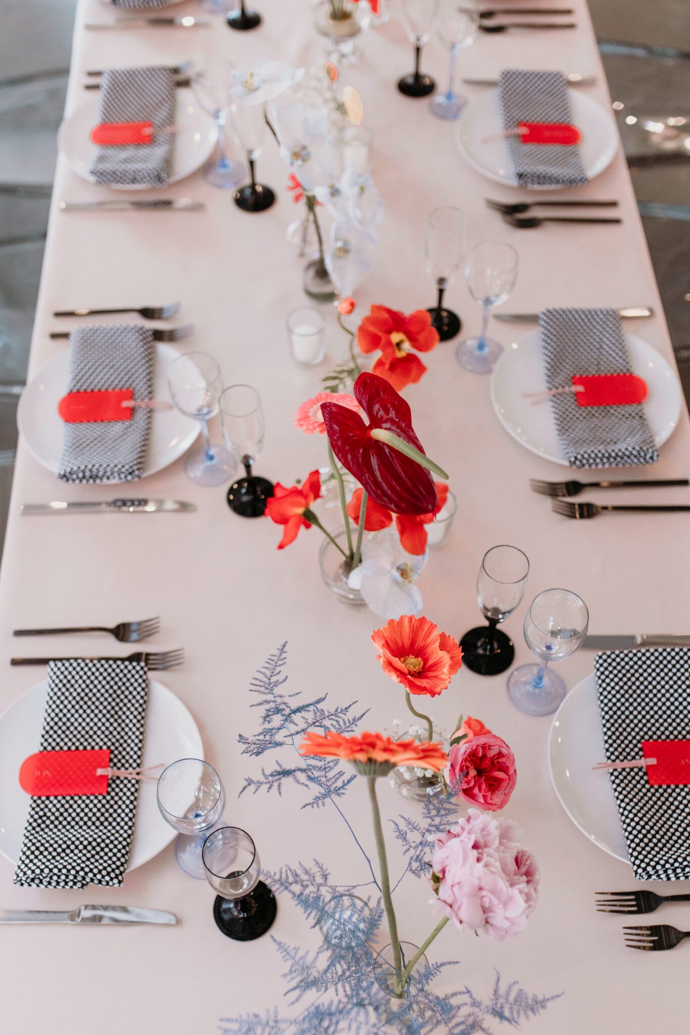 A contemporary wedding tablescape blending modern and retro elements: light pink tablecloth, grey ghost chairs, black acrylic place cards, patterned napkins, retro-inspired table numbers, and ikebana floral arrangements with anthurium flowers.