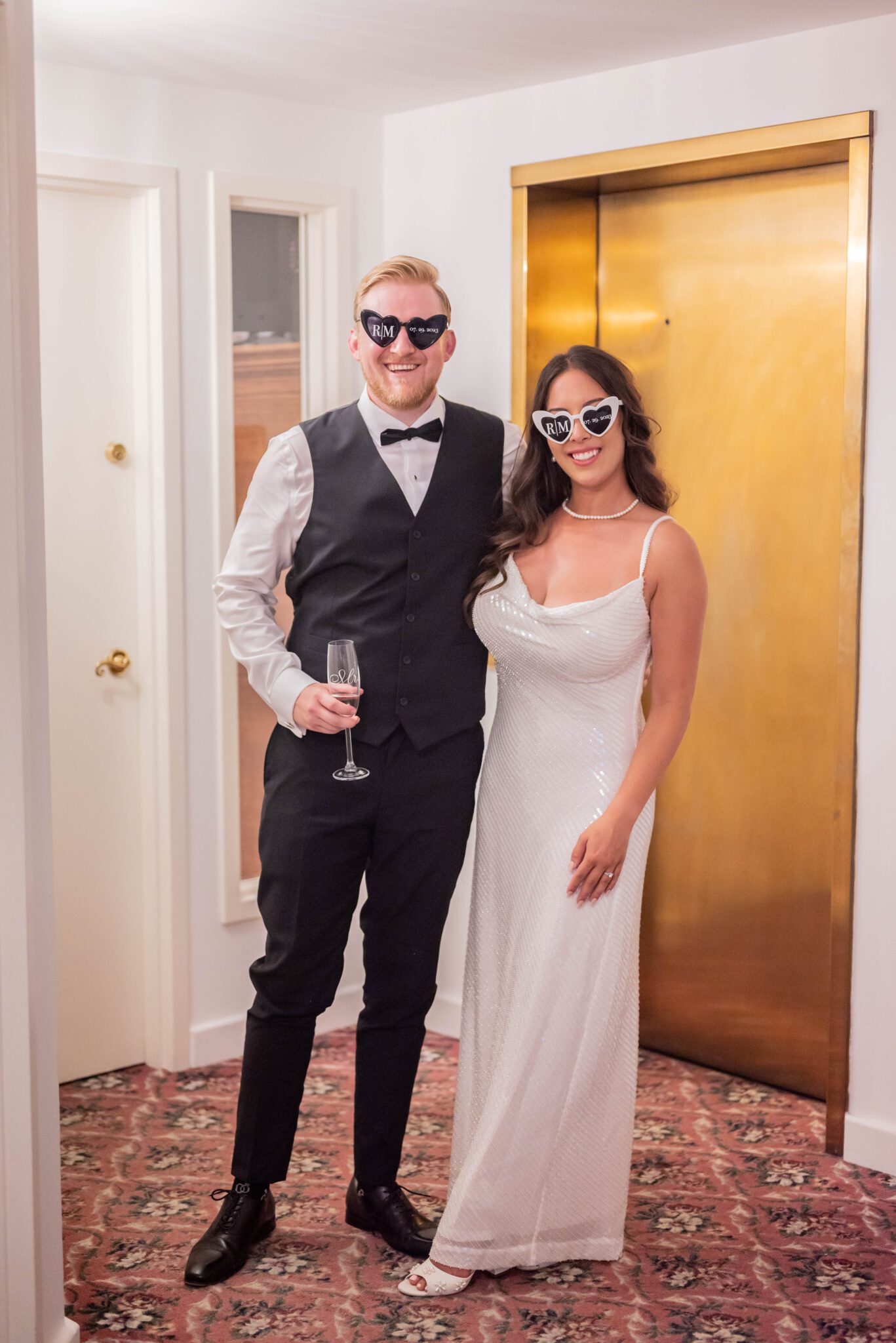 Portrait of bride and groom wearing their second outfits for their reception, featuring bride in sequin gown and heart-shaped sunglasses.