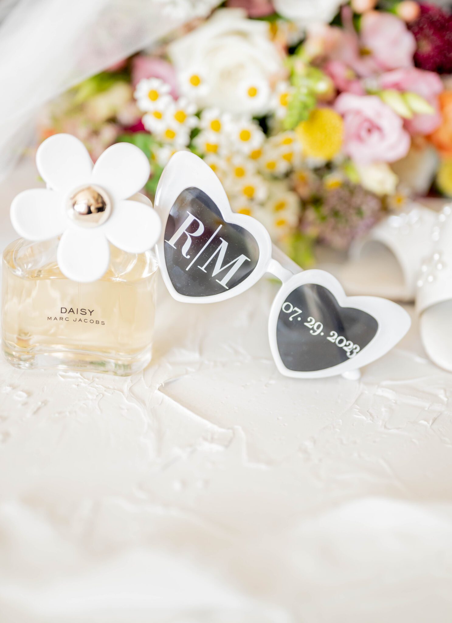 Flatlay photo of bride's custom heart-shaped sunglasses and the bride's Marc Jacobs perfume bottle, with colourful florals in the background. 