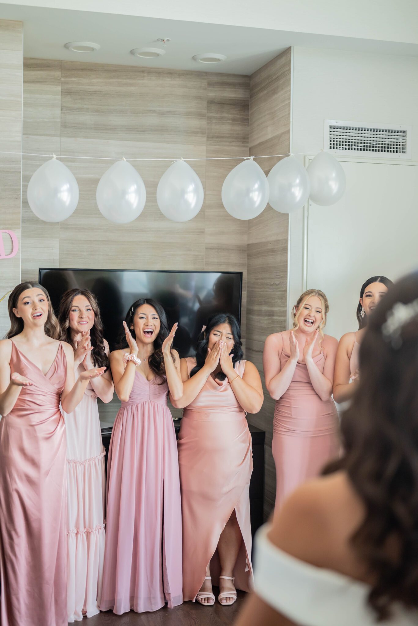 Bridesmaid first look reaction to bride in her elegant gown, bridesmaids in different shades of pink gowns, in getting ready suite. 