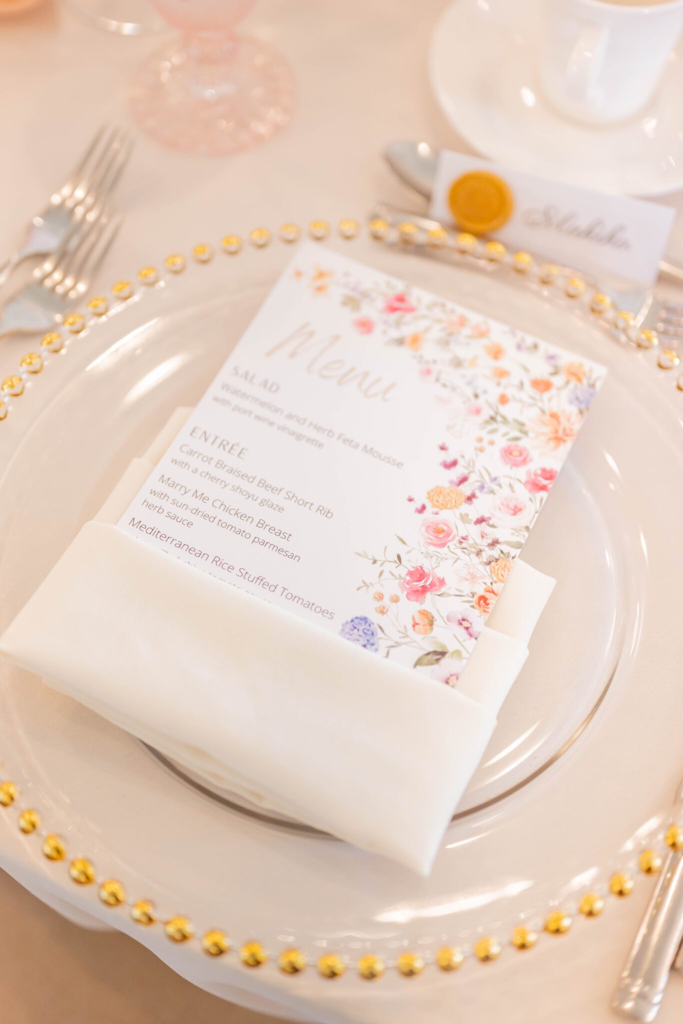 Close up detailed photo of custom wedding menus, featuring a floral-printed design on top of gold-rimmed charger plates. 