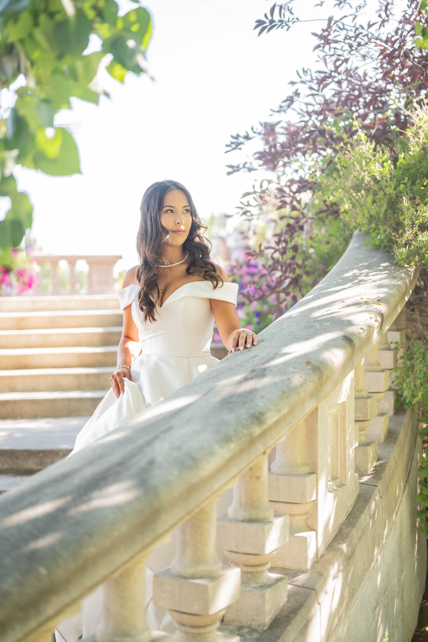 Bride walks down the elegant staircase at Spruce Meadows, surrounded by greenery and foliage, wearing chic gown with off the shoulder sleeves and a sweetheart neckline. 