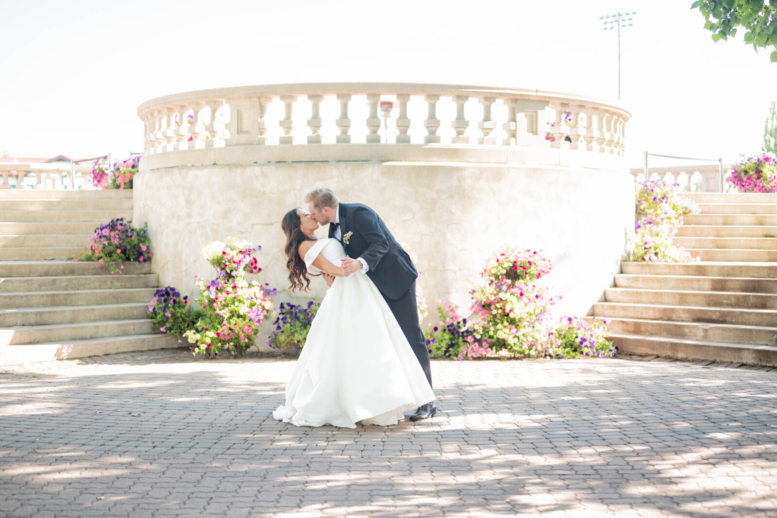 Bride and groom share a dip kiss in front of the elegant staircase and vintage architecture at Spruce Meadows, surrounded by colourful flowers and greenery.