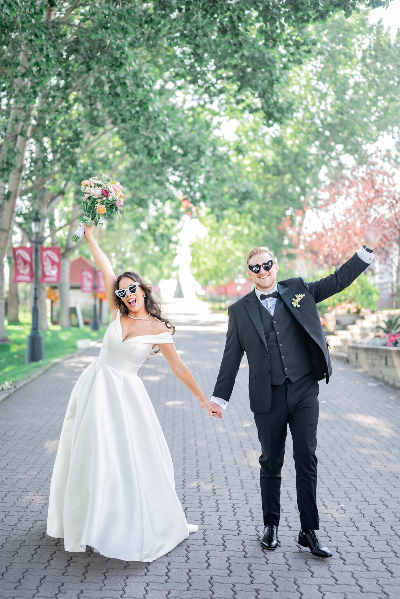 Couple raises their arms in celebration at their whimsical garden wedding at Spruce Meadows, featuring custom heart shaped sunglasses and a colourful bridal bouquet. 