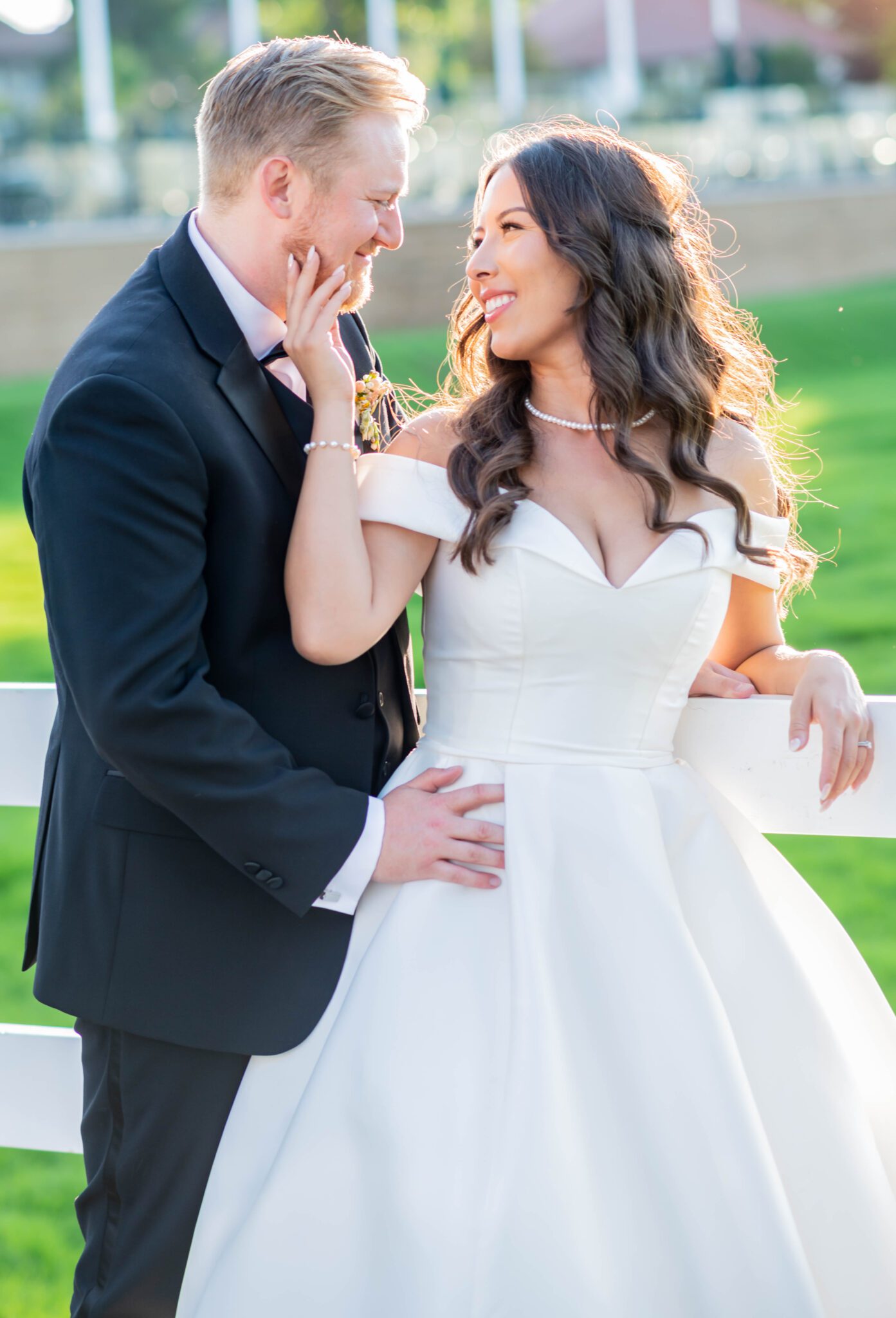 Portrait of bride and groom at Spruce Meadows, leaning against a fence, embracing each other, bride in elegant white gown with off the shoulder sleeves and a sweetheart neckline.