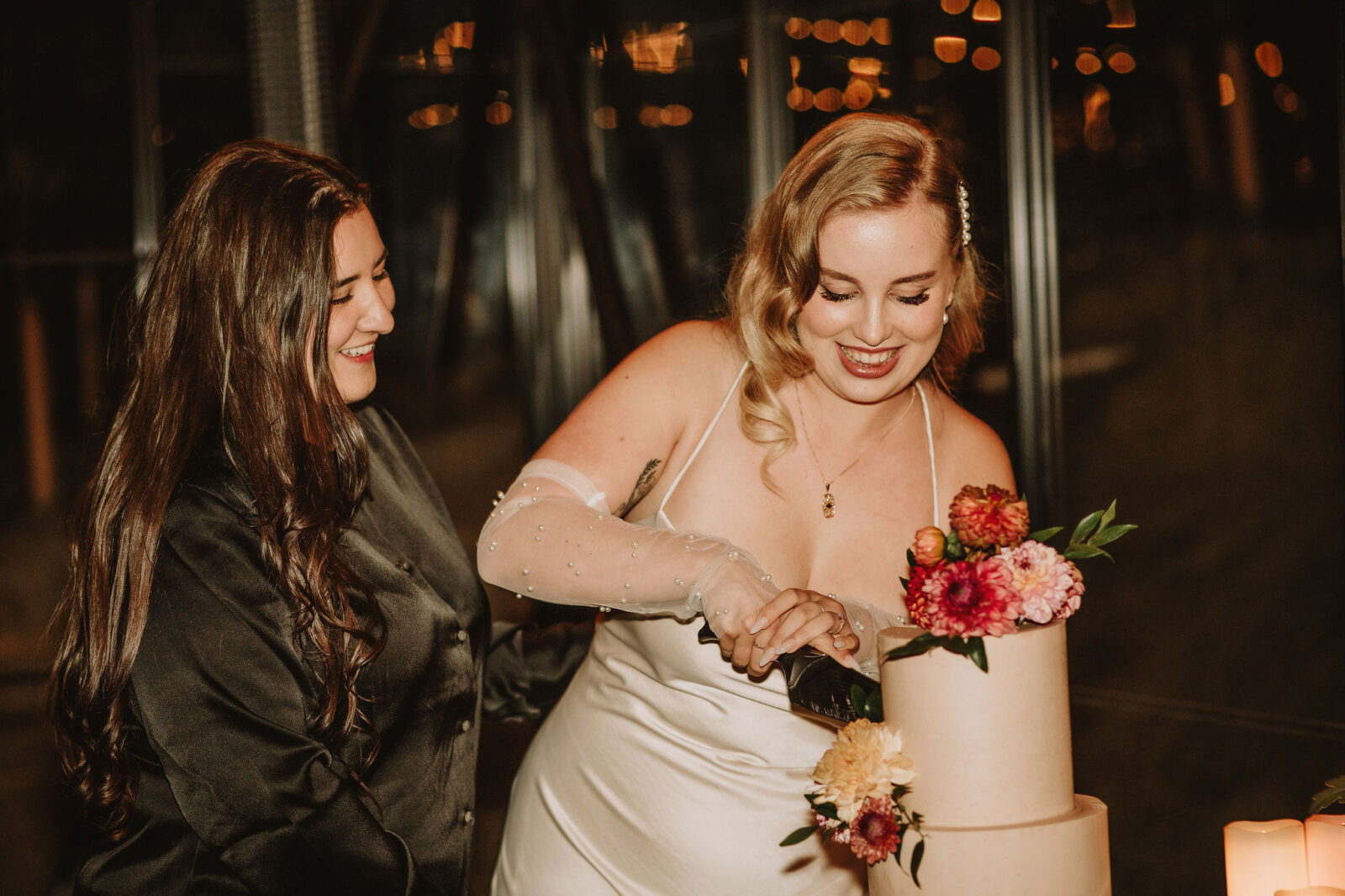 Portrait of couple cutting their wedding cake, topped with colourful florals, featuring bride in silk mini dress and sheer gloves. 