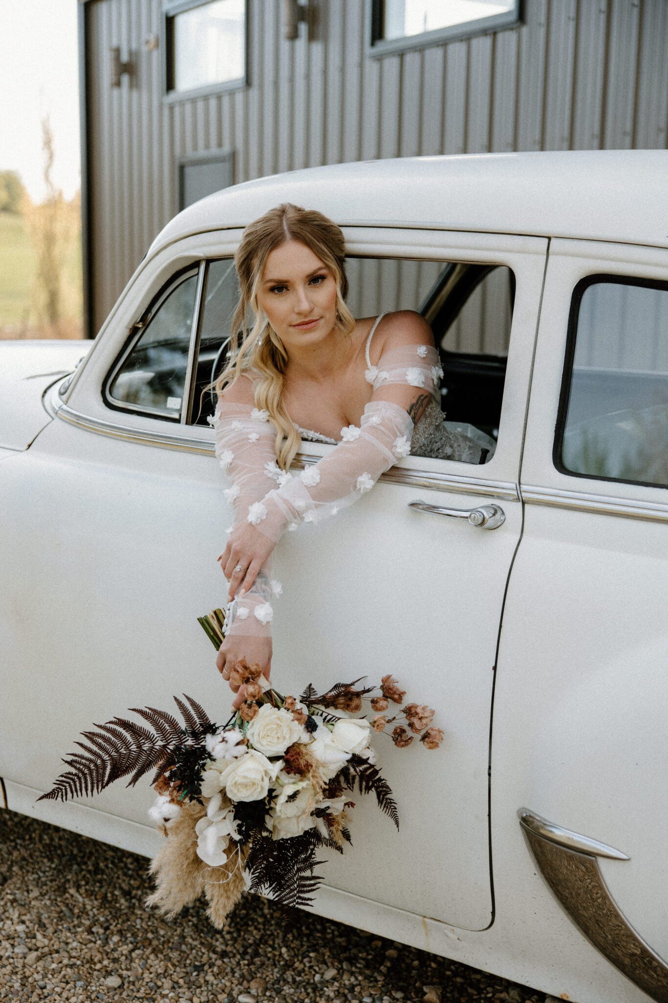 Portrait of bride sitting in the vintage getaway car at 52 North Venue, holding the bridal bouquet with rich textures and tones, wearing a floral detailed bridal gown with her hair in an elegant ponytail updo. 