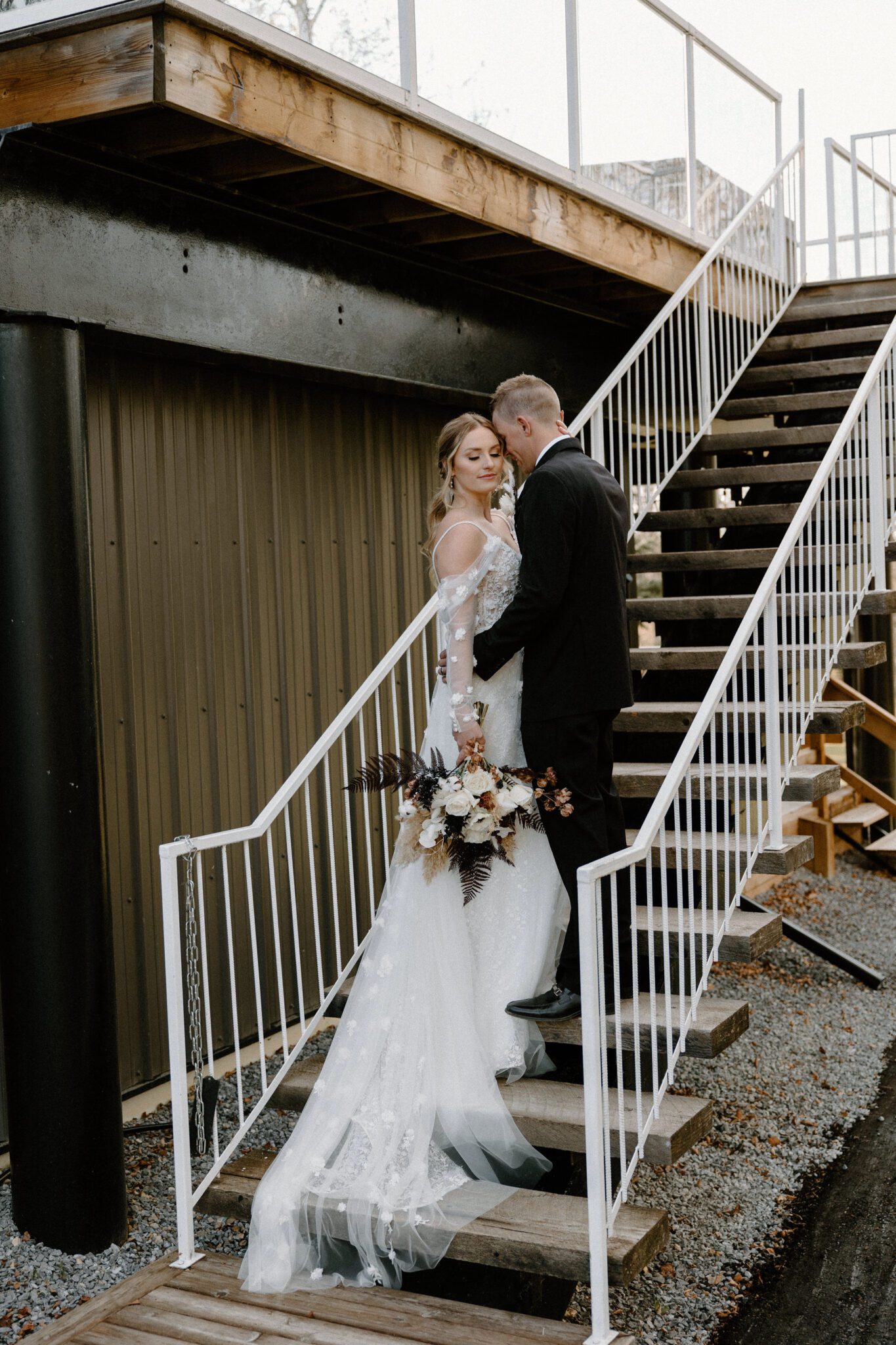 Elegant portrait of bride and groom at 52 North Venue on a staircase, with bride's floral-detailed dress draping down, warm and moody wedding inspiration.