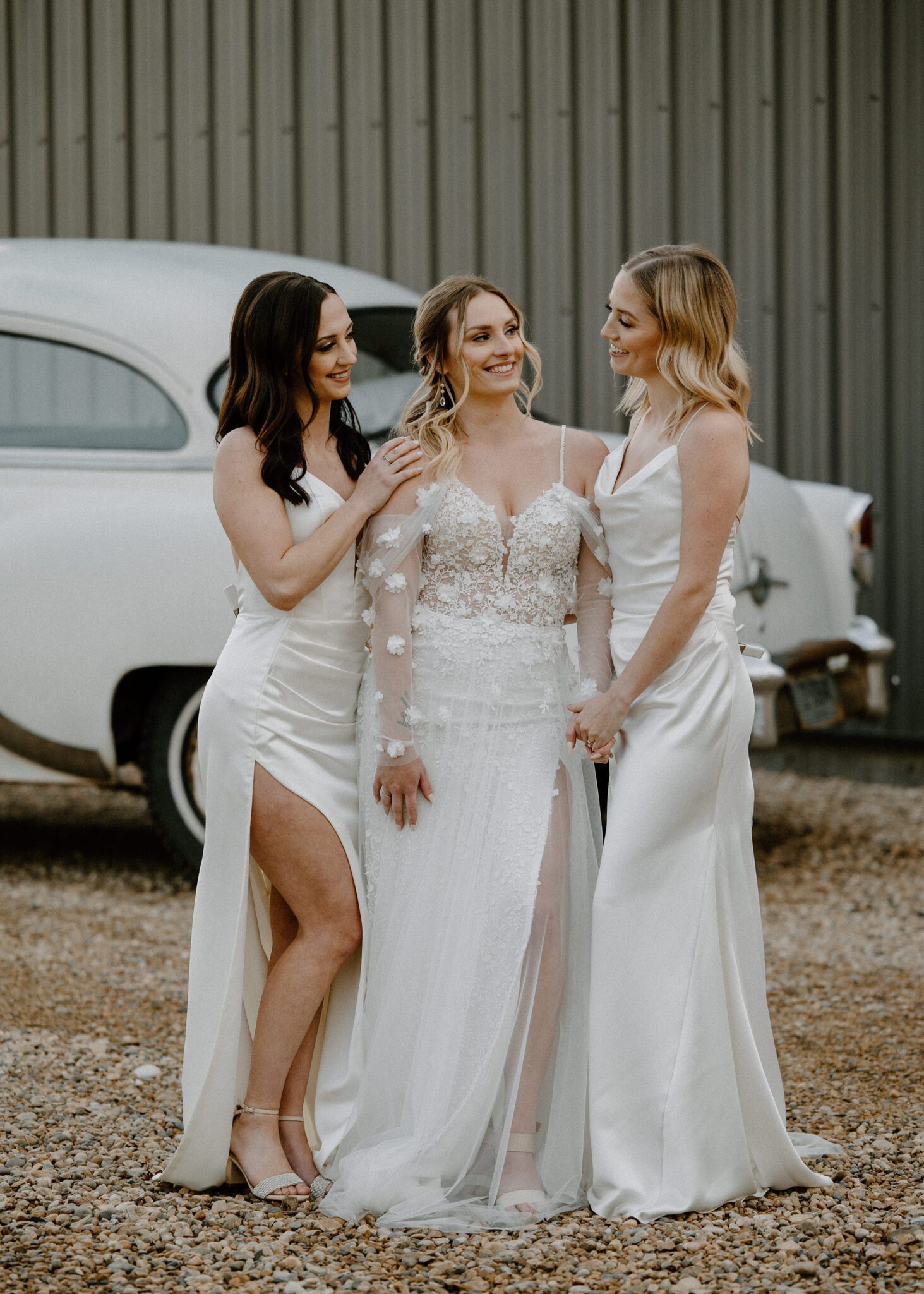 Bride and her bridesmaids standing in front of cream-coloured vintage getaway car, in white gowns, warm and moody wedding inspiration at the 52 North Venue.