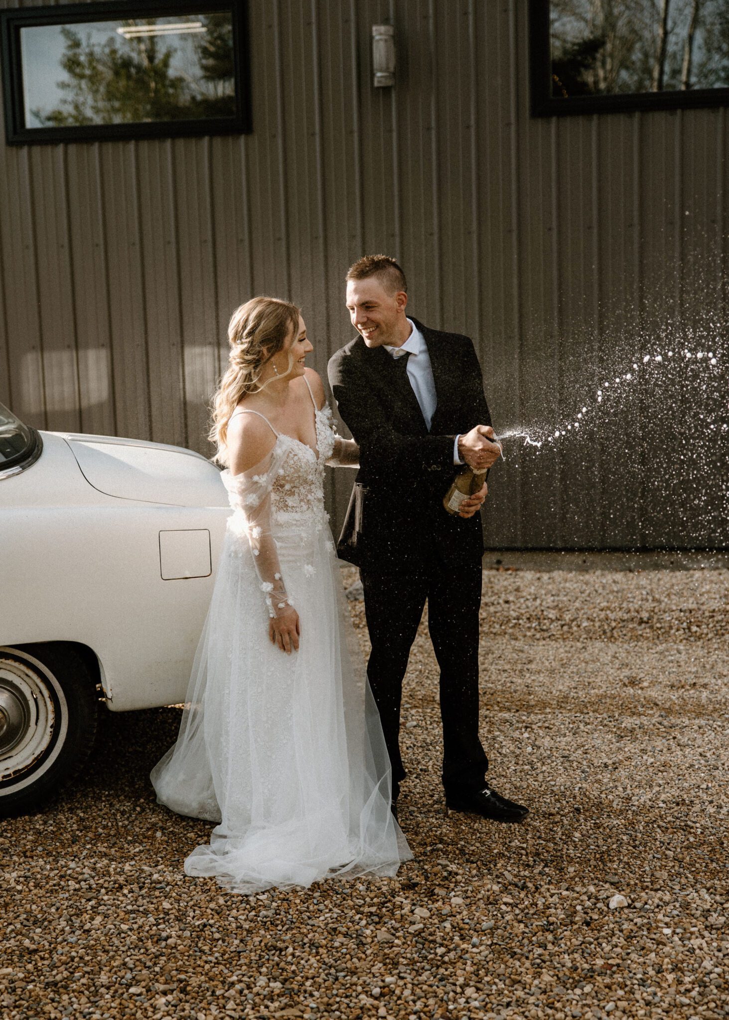 Bride and groom popping a champagne bottle in celebration beside their cream-coloured vintage getaway car, warm and moody wedding inspiration at 52 North Venue. 