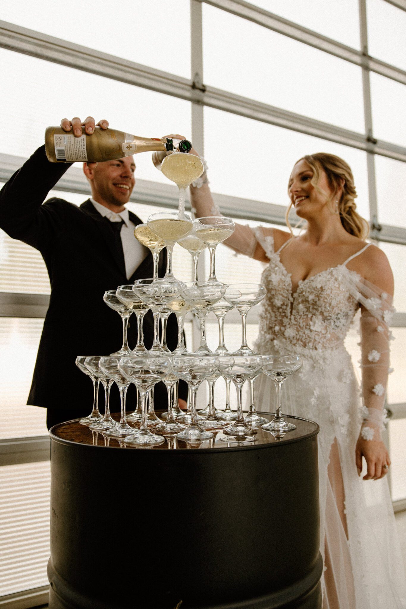 Bride and groom celebrating with a champagne tower at 52 North Venue, bride in floral-detailed tulle gown with sheer sleeves, warm and moody wedding inspiration. 