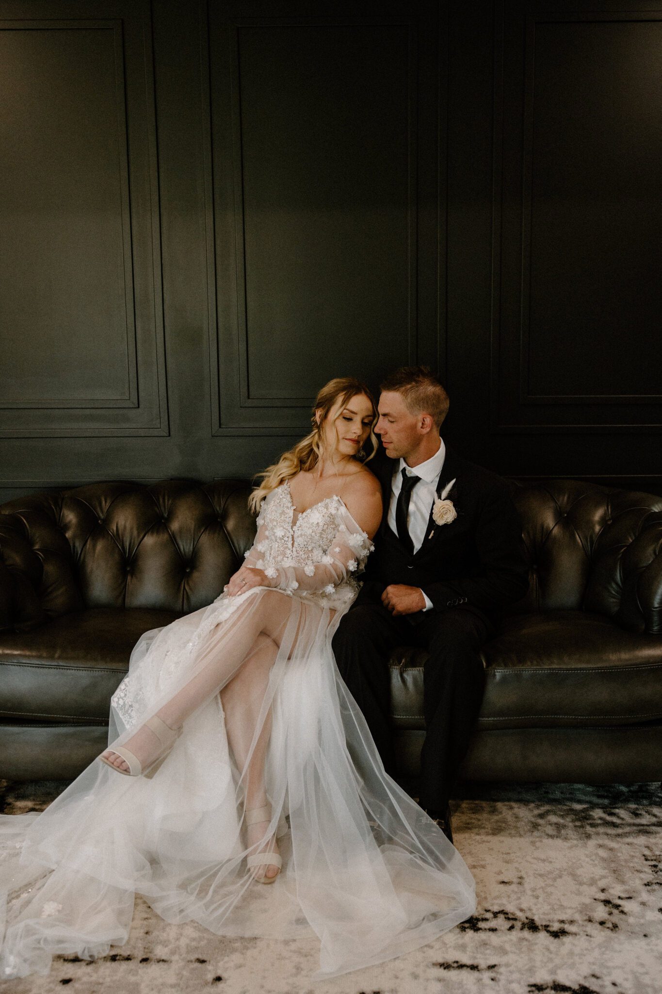 Indoor portrait of bride and groom sitting on vintage couch at 52 North Venue, bride in floral-detailed tulle gown, warm and moody wedding inspiration. 