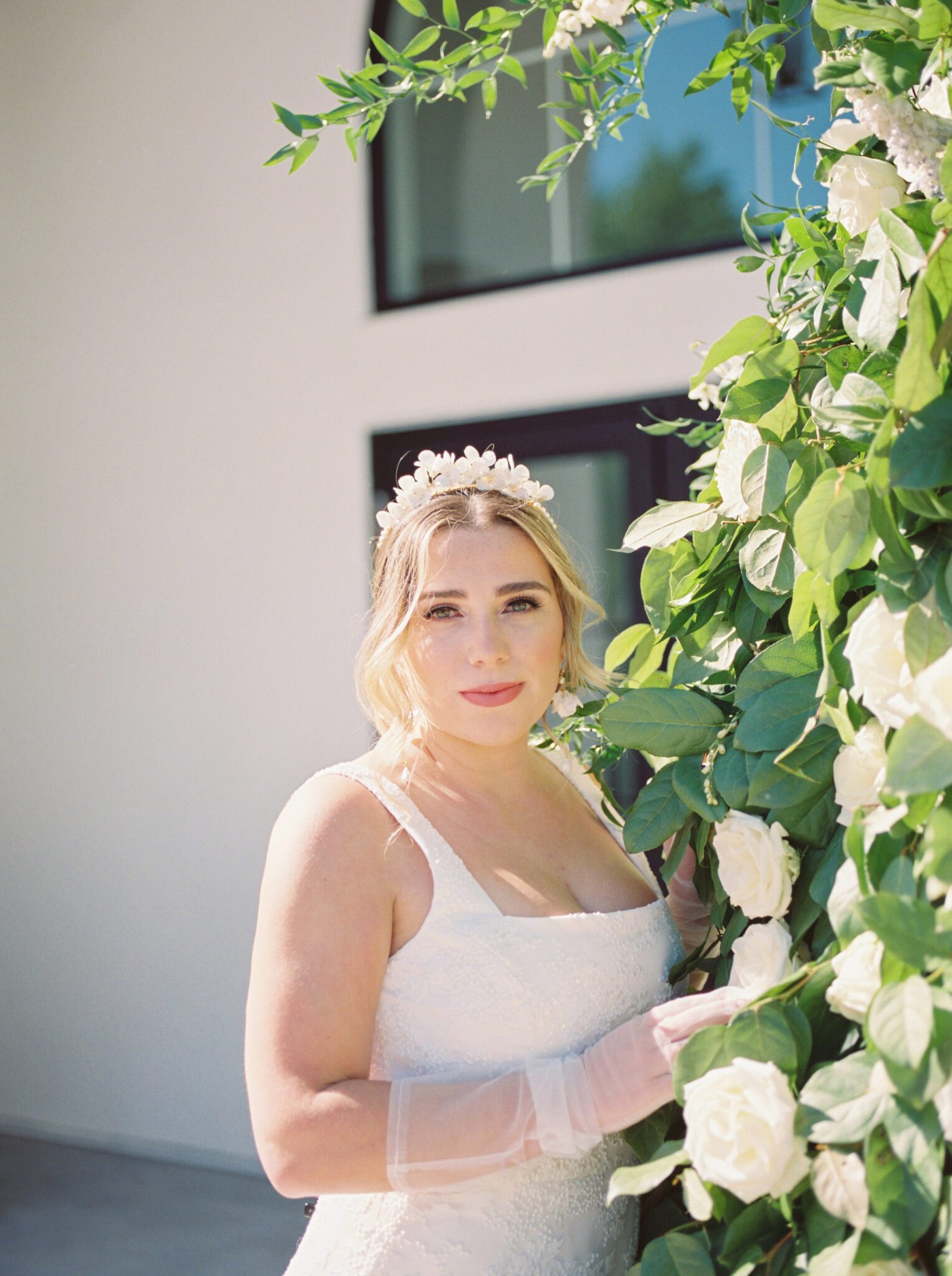 Bride standing in front of lush ivory, white and green floral installation wearing sophisticated yet whimsical bridal gown and jewelry. Outdoor summer wedding inspiration.