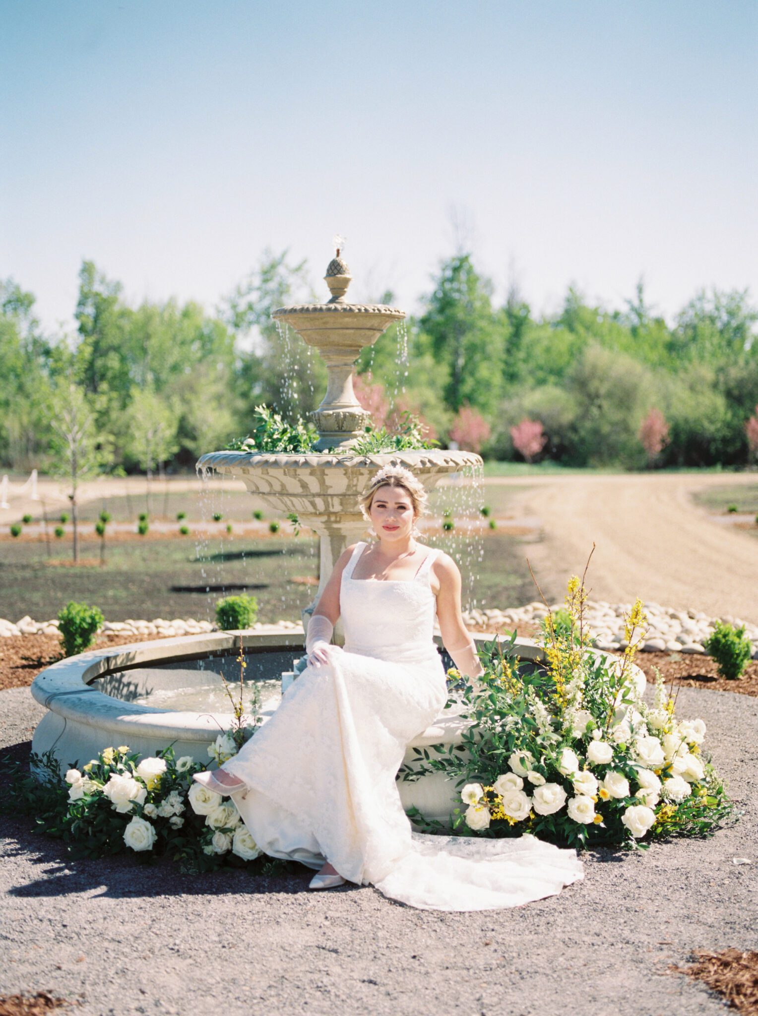 Gorgeous bride sitting on the elegant water fountain at Sparrow Lane Events, surrounded by lush pale yellow and ivory florals by Lovella Lifestyle.